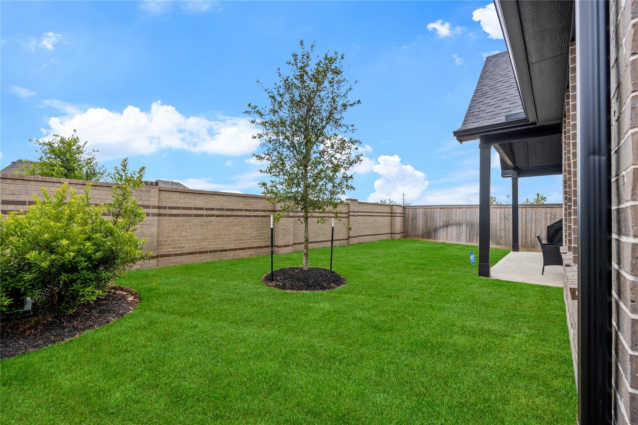 Fenced backyard with a broad lawn, young tree, brick privacy wall, and covered patio.