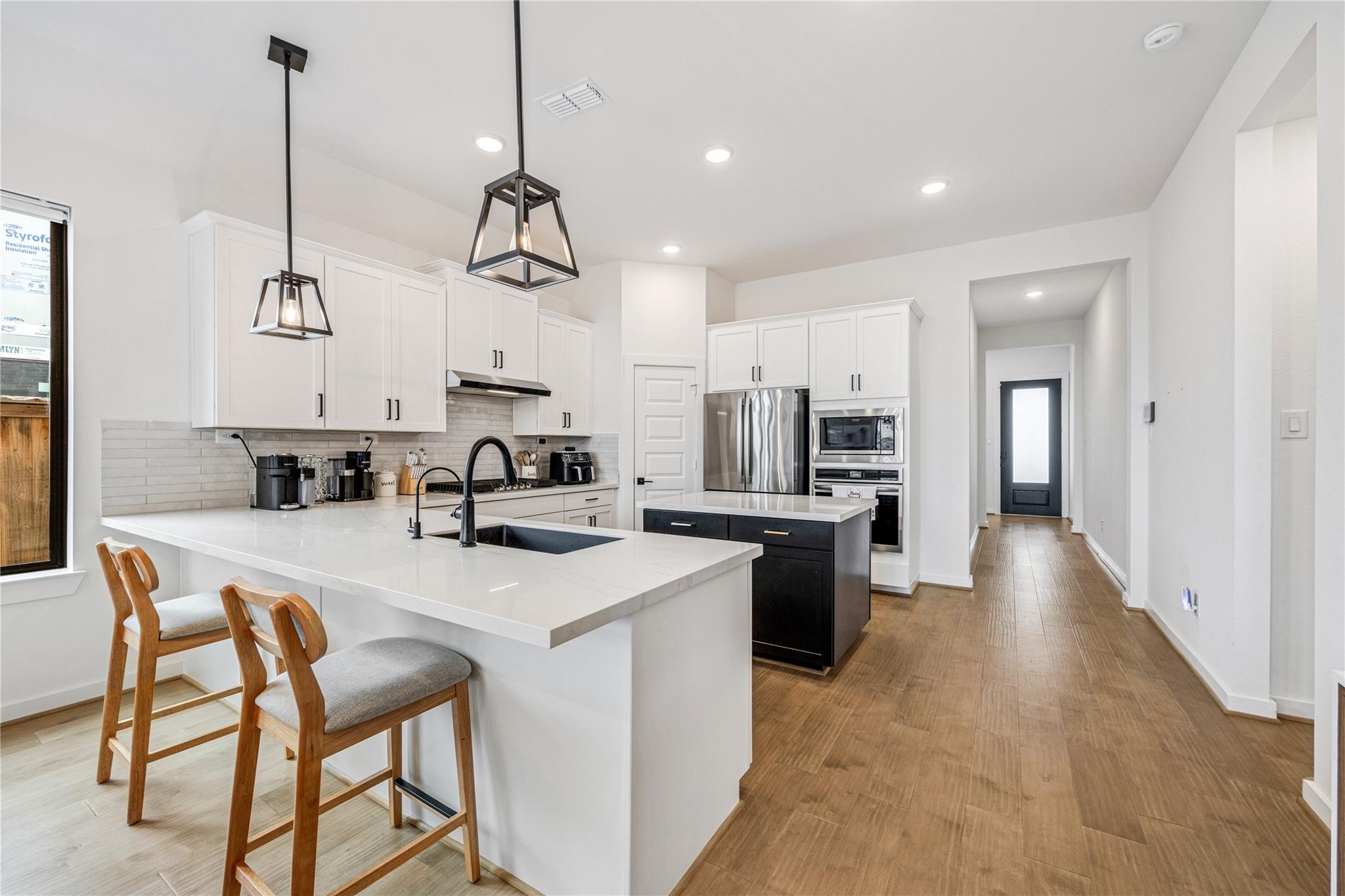 Modern white kitchen with a large island, wood bar stools, black pendant lights, and stainless steel appliances.