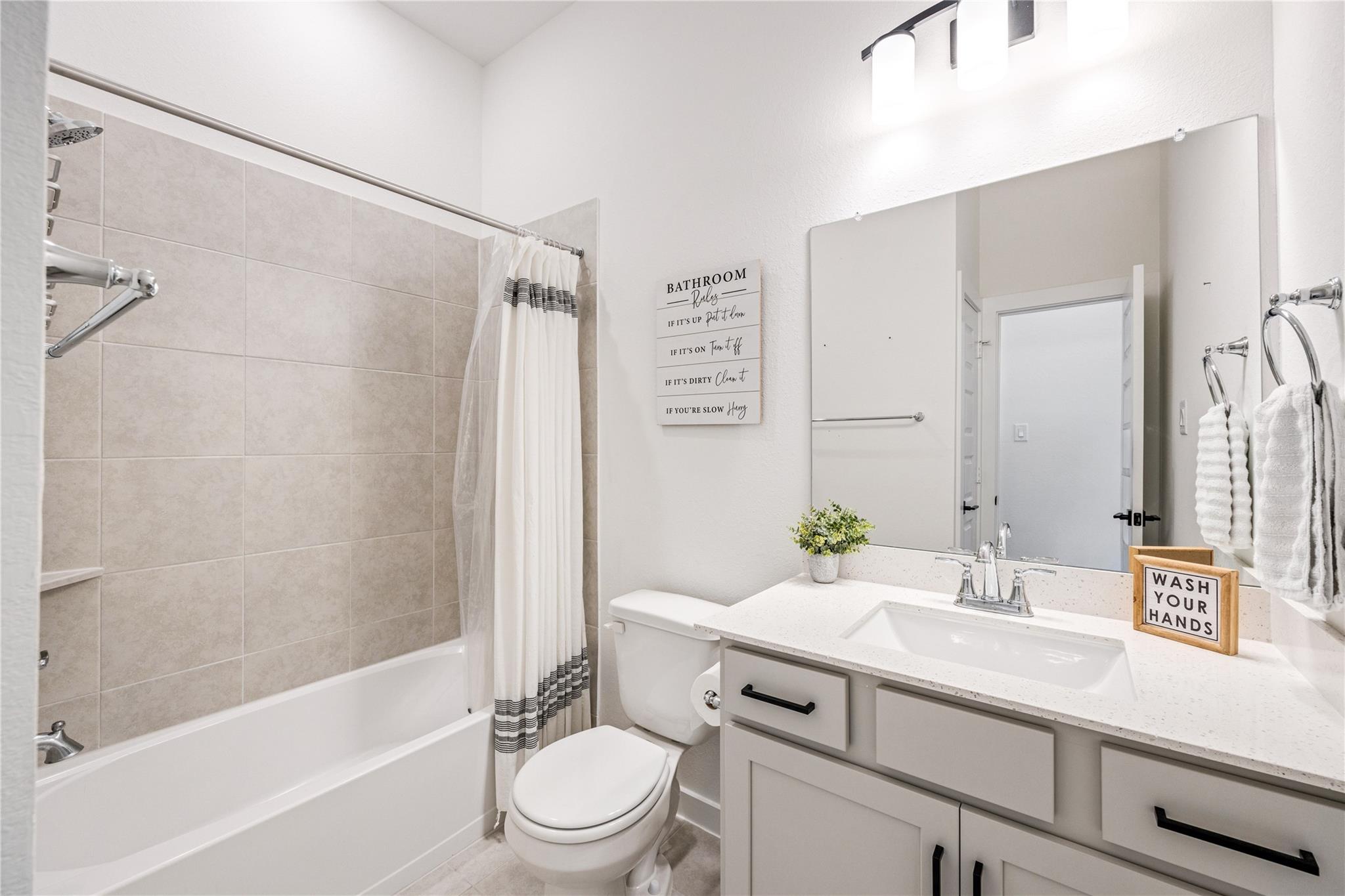 Bathroom with a tiled tub and shower, striped curtain, toilet, and gray vanity with a large mirror.