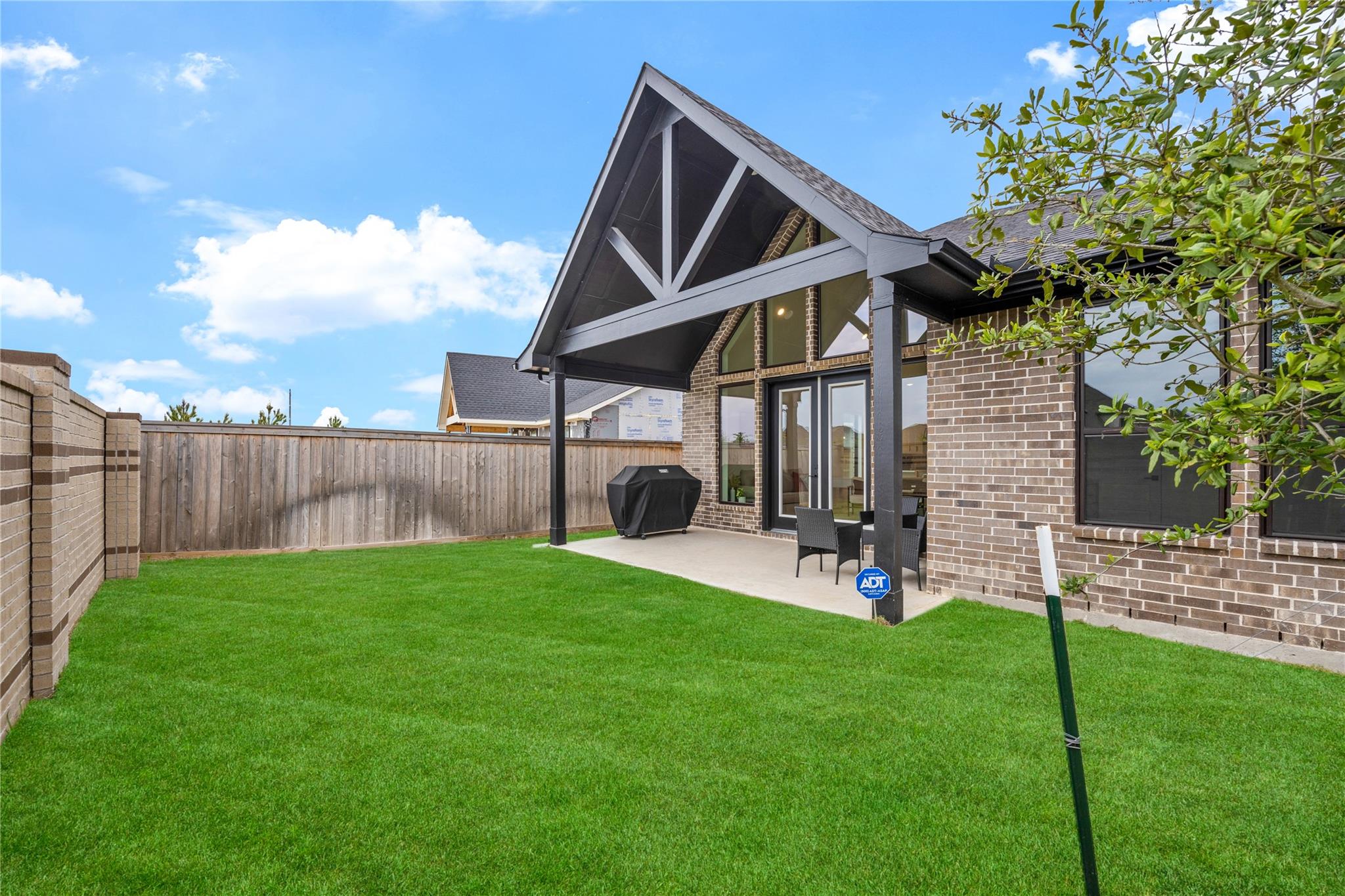 Brick home’s covered patio and peaked gable overlook a fenced backyard with a wide green lawn.