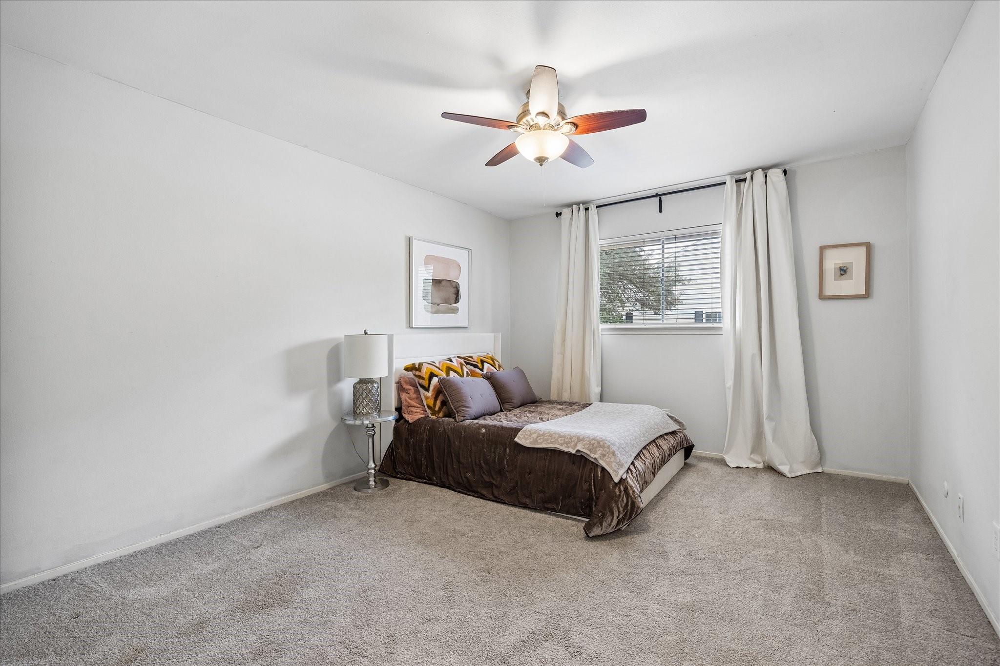 Furnished bedroom with a brown-covered bed, gray carpet, curtained window, and ceiling fan.