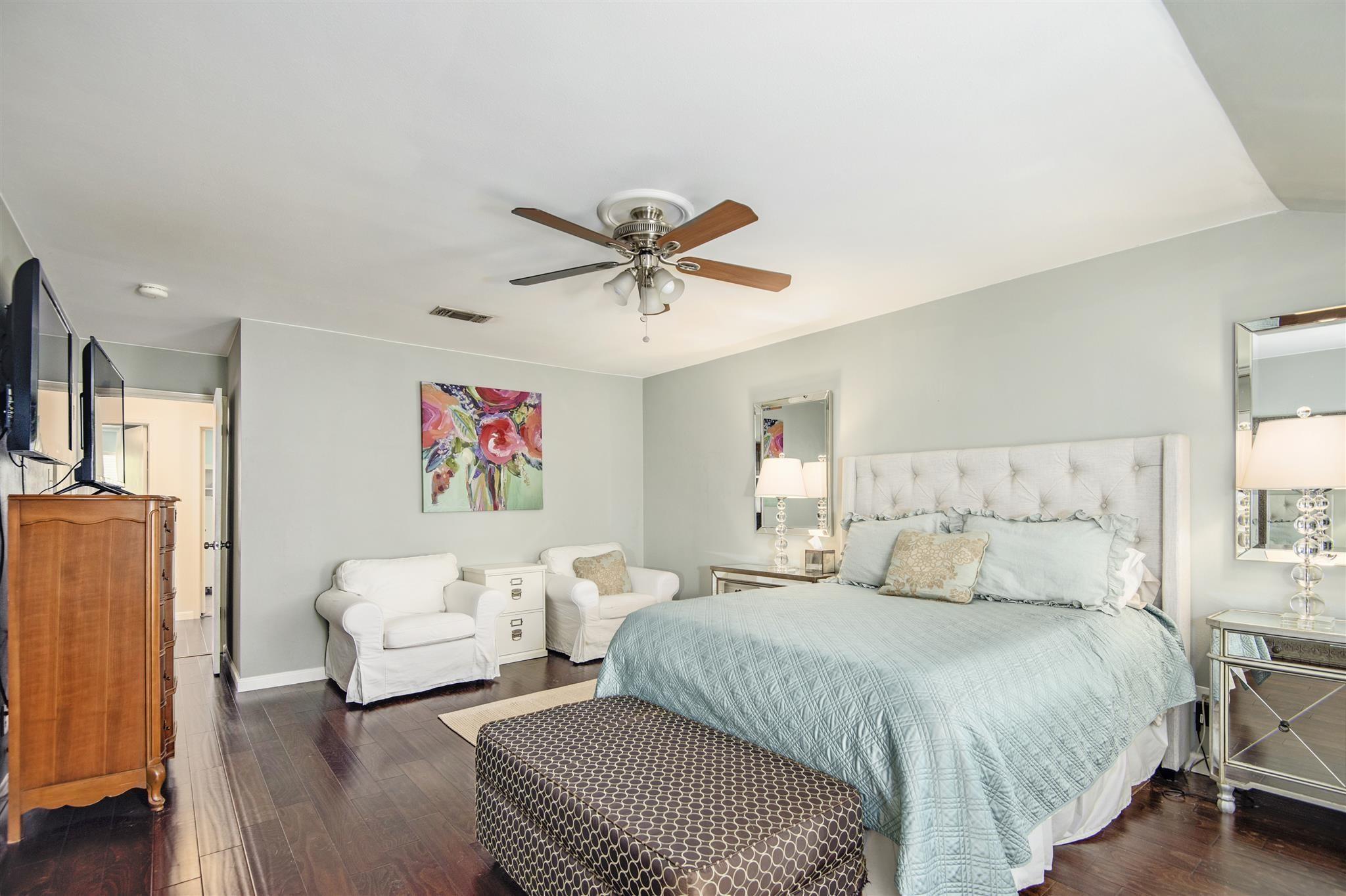 Furnished bedroom with a pale blue bedspread, tufted headboard, ceiling fan, and two white armchairs.