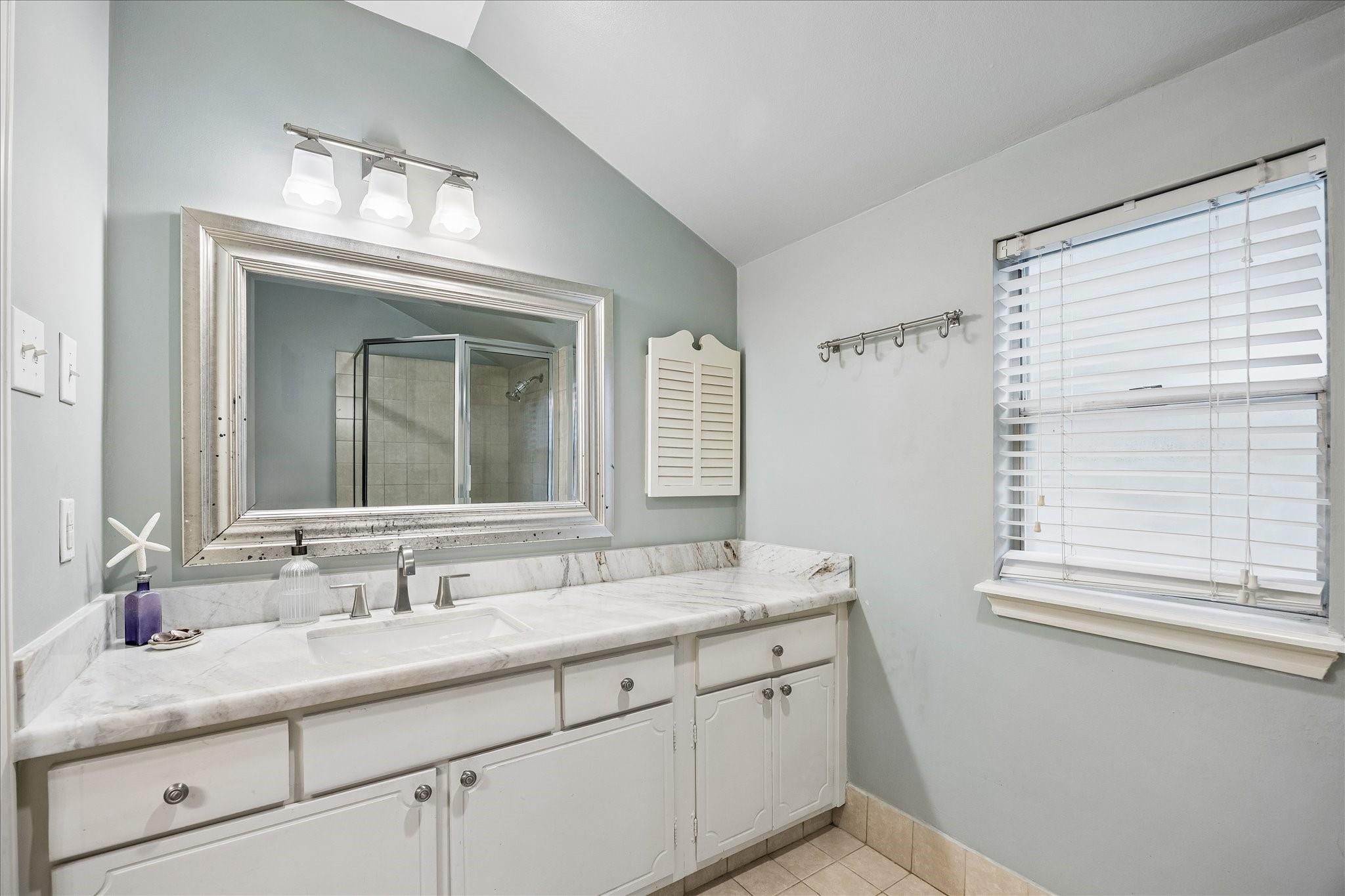 Bathroom with a long marble-topped vanity, large framed mirror, and window with white blinds.