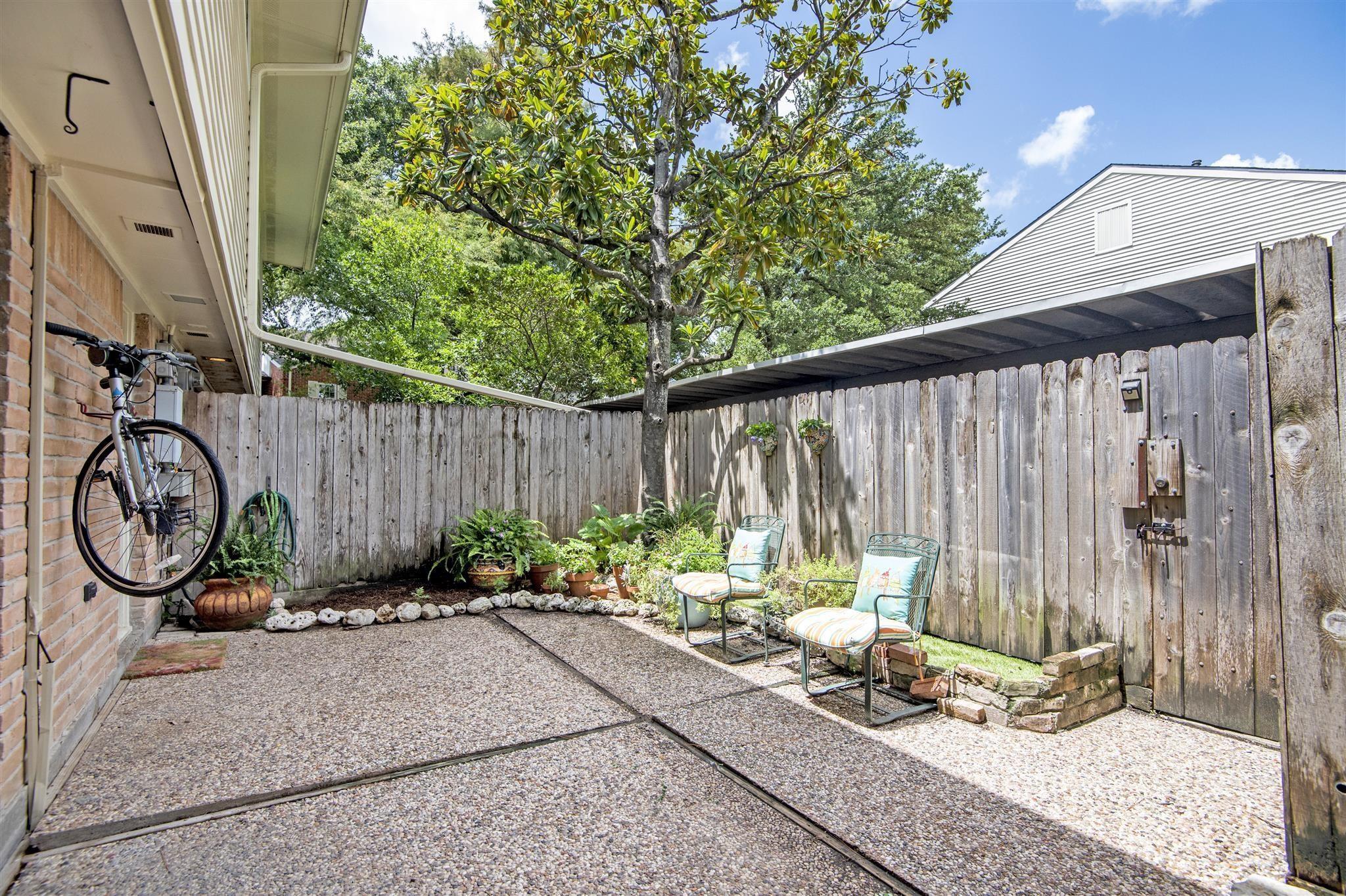 Fenced pebble patio with two chairs, potted plants, and a mature shade tree.