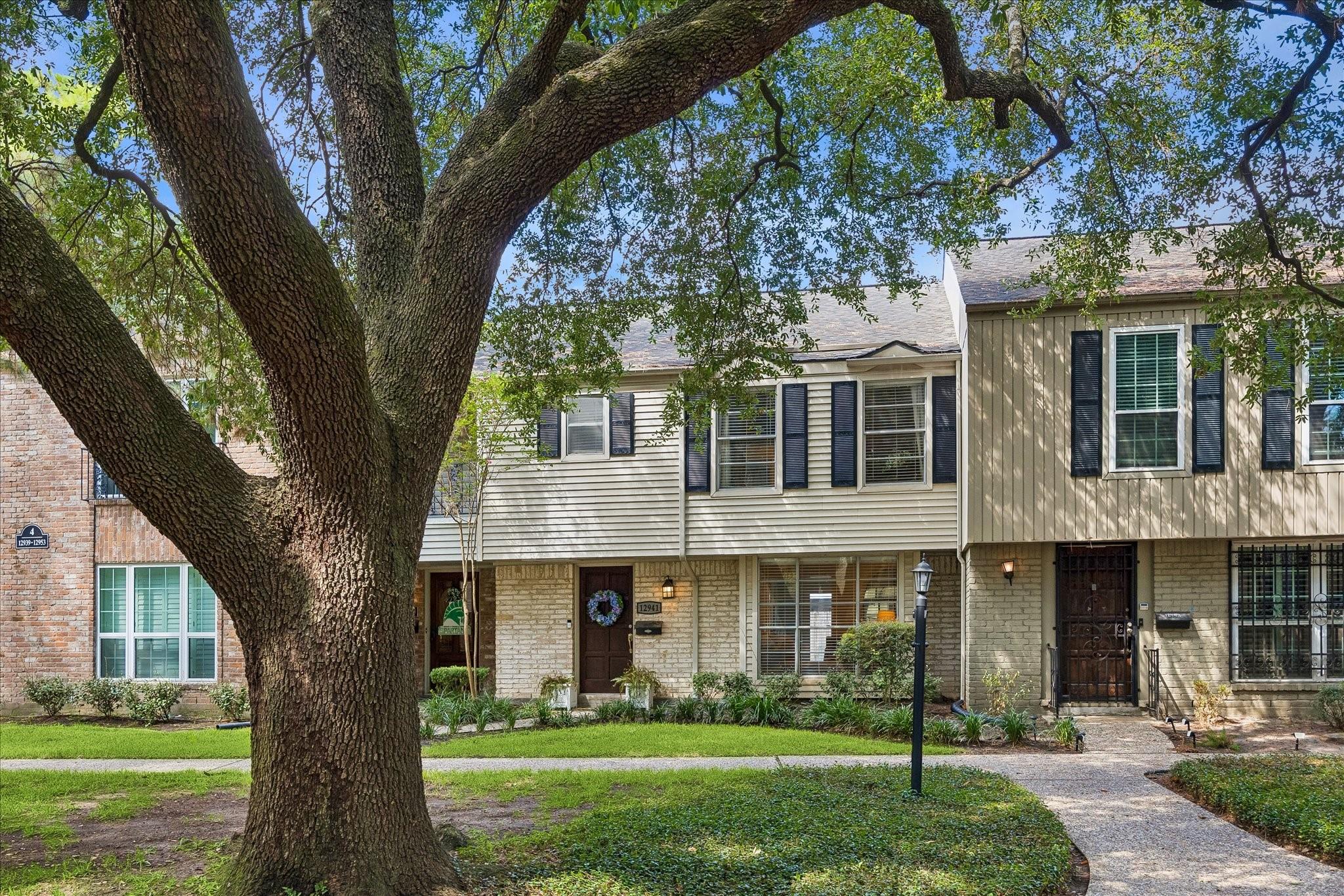 Attached two-story townhomes behind a large shade tree, with a landscaped walkway and front entrances.