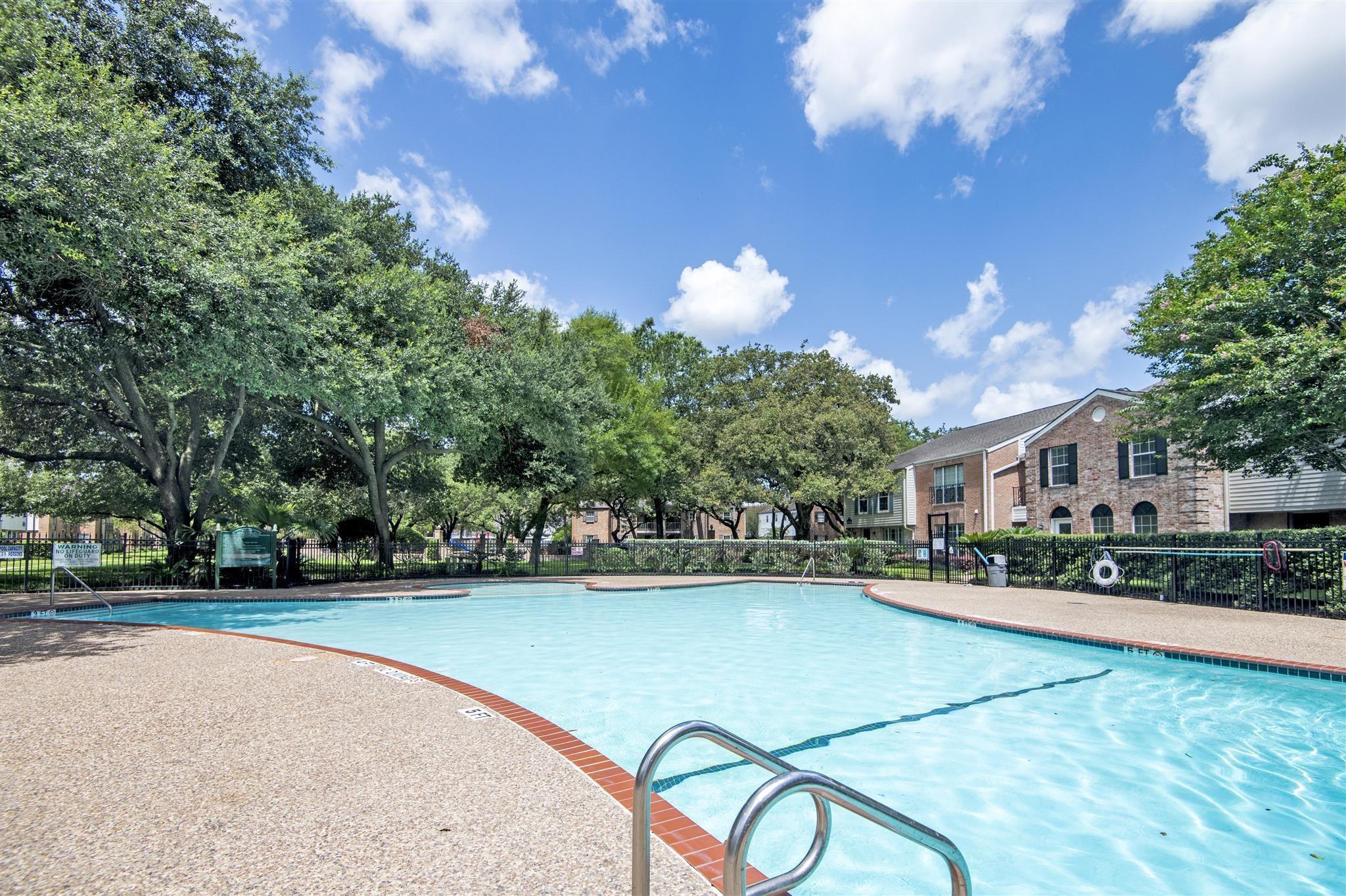 Community swimming pool with curved brick edging, mature trees, and brick apartment buildings.