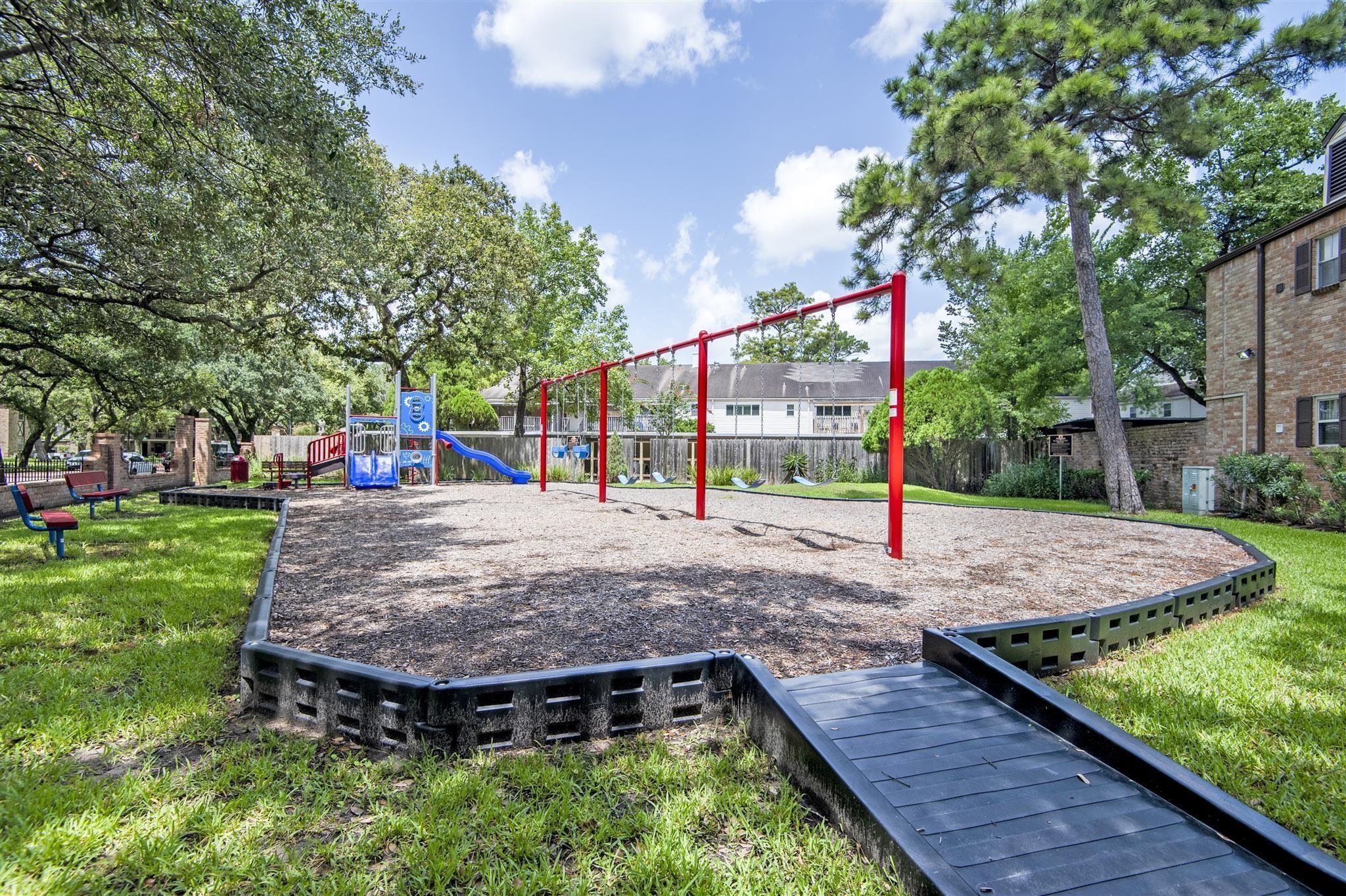 Community playground with red swings, a blue slide, benches, and mature trees.