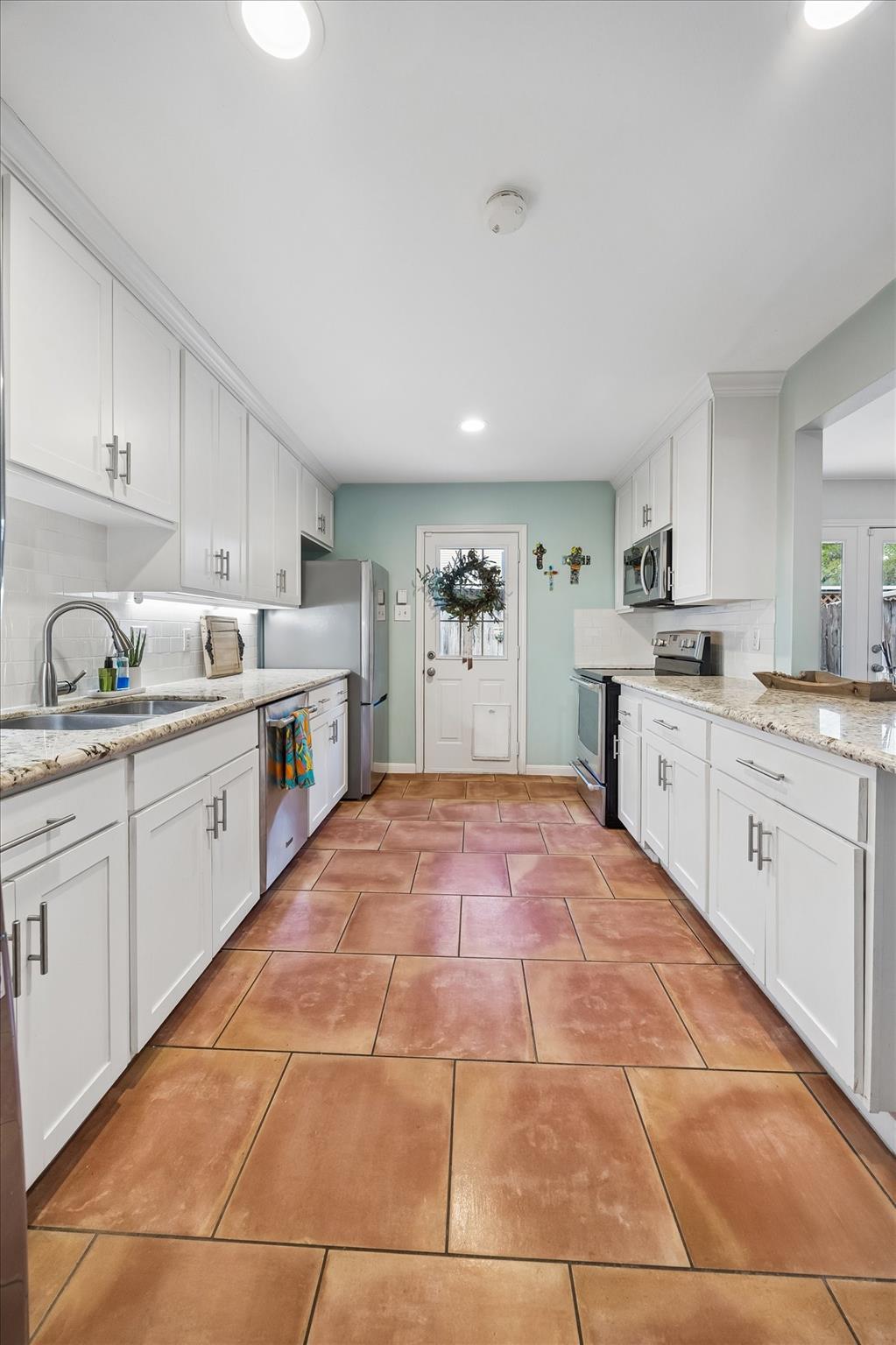 Galley kitchen with white cabinets, granite counters, stainless appliances, and large terracotta floor tiles.