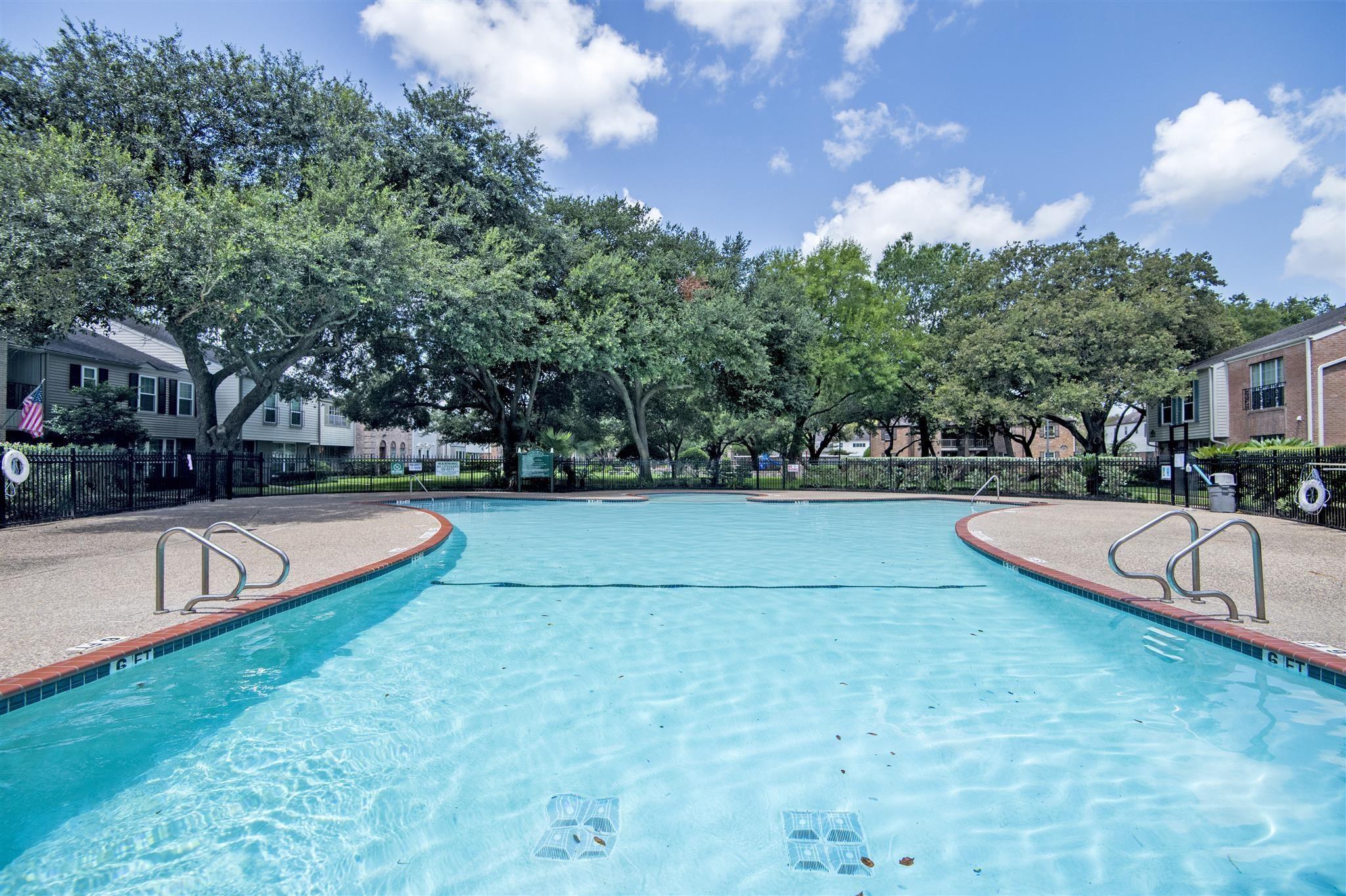 Community swimming pool with metal ladders, a black fence, and mature trees.