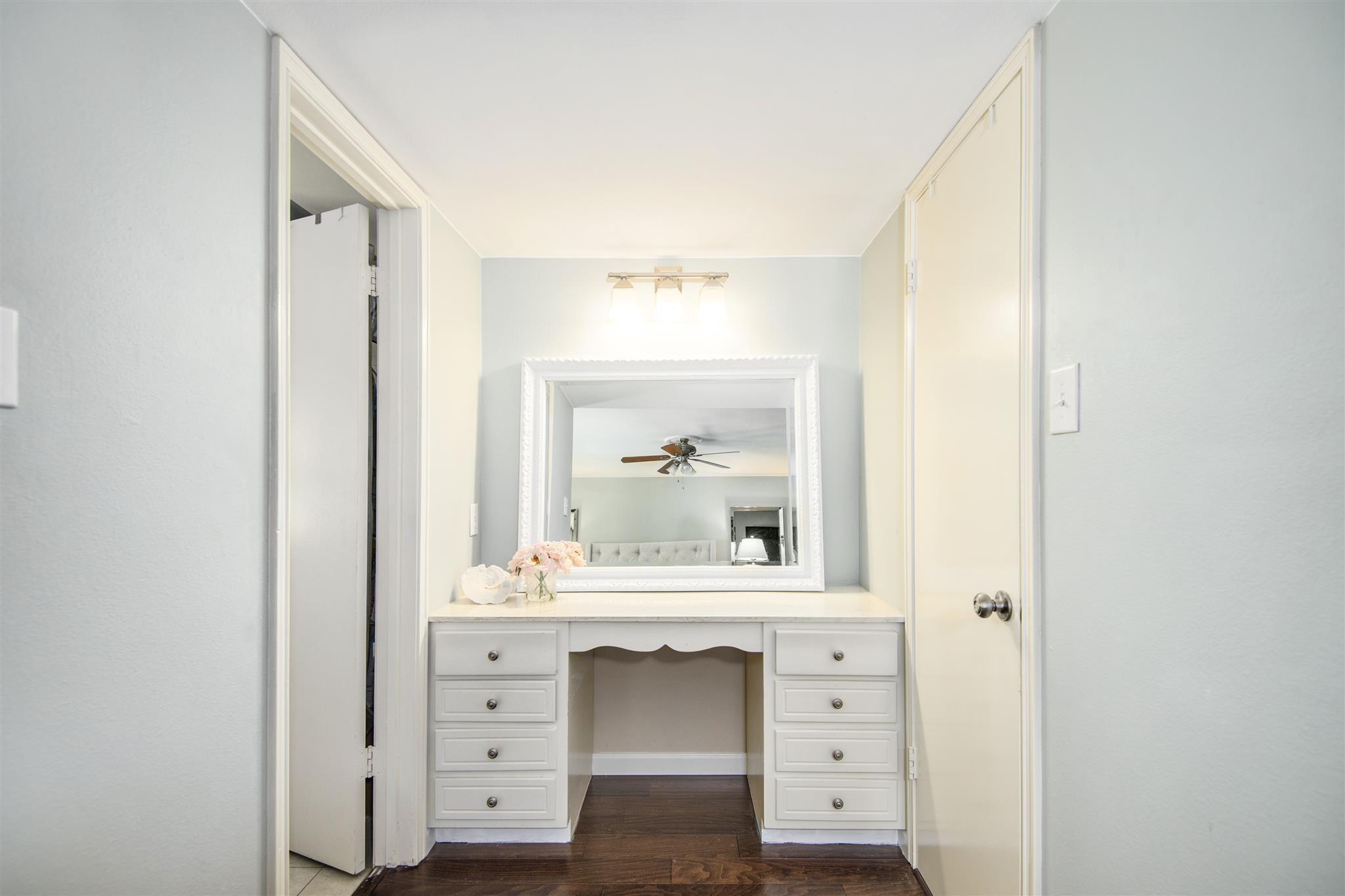 Built-in white makeup vanity with a large mirror and drawers, opening toward a bedroom.