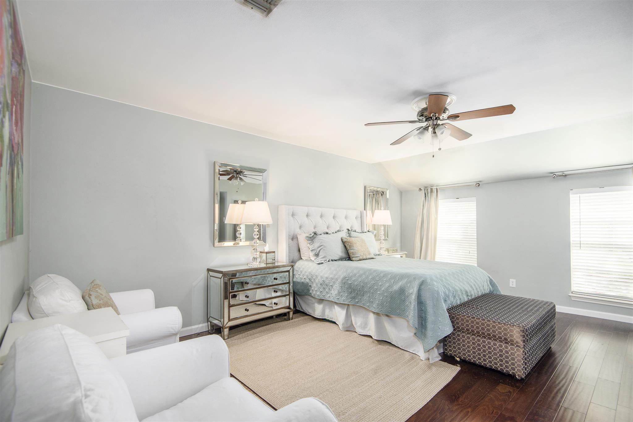 Bedroom with a light blue quilted bed, tufted headboard, white seating area, and dark wood floors.