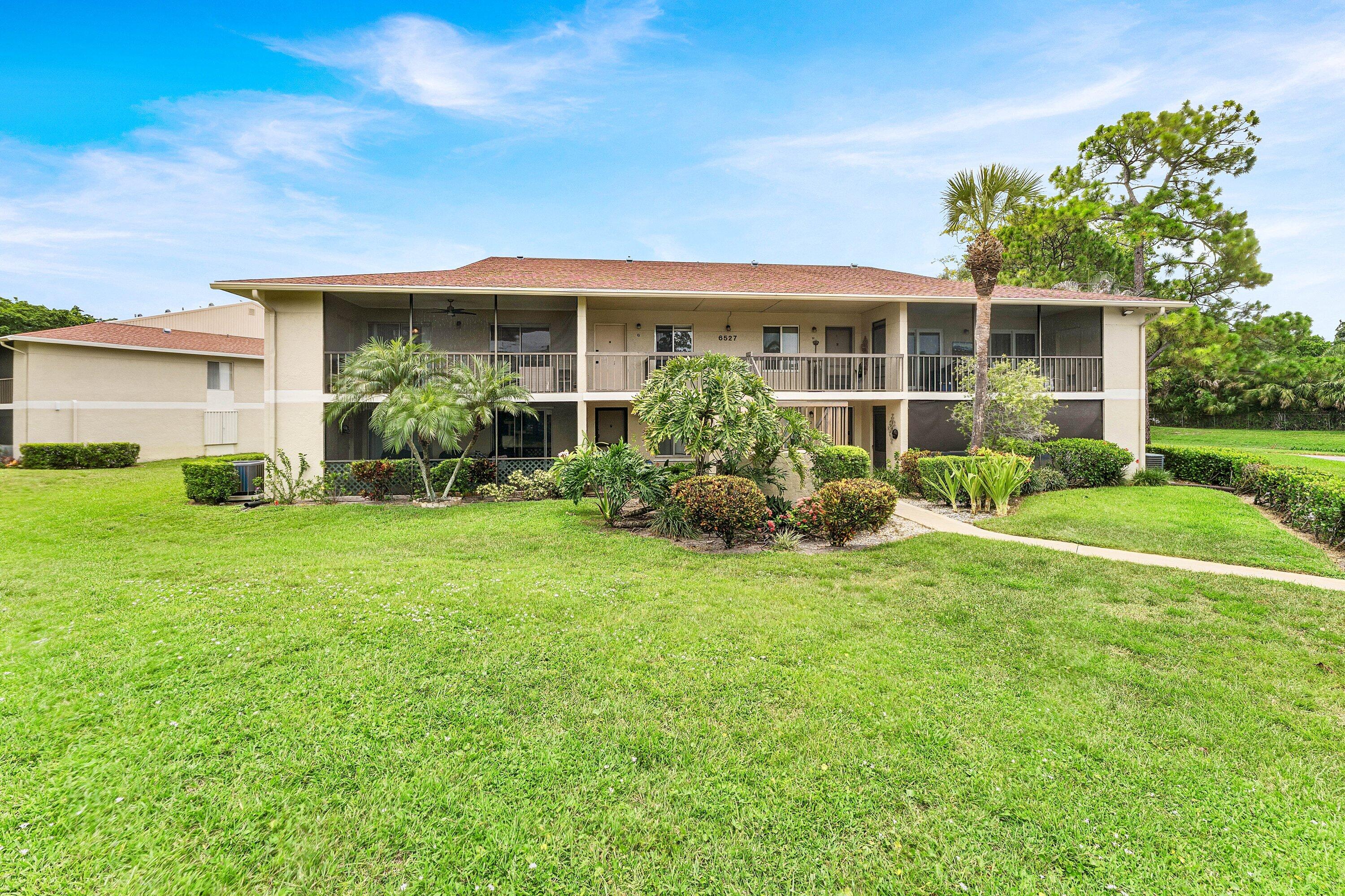 Two-story beige condominium building with screened balconies, tropical landscaping, and a broad green lawn.