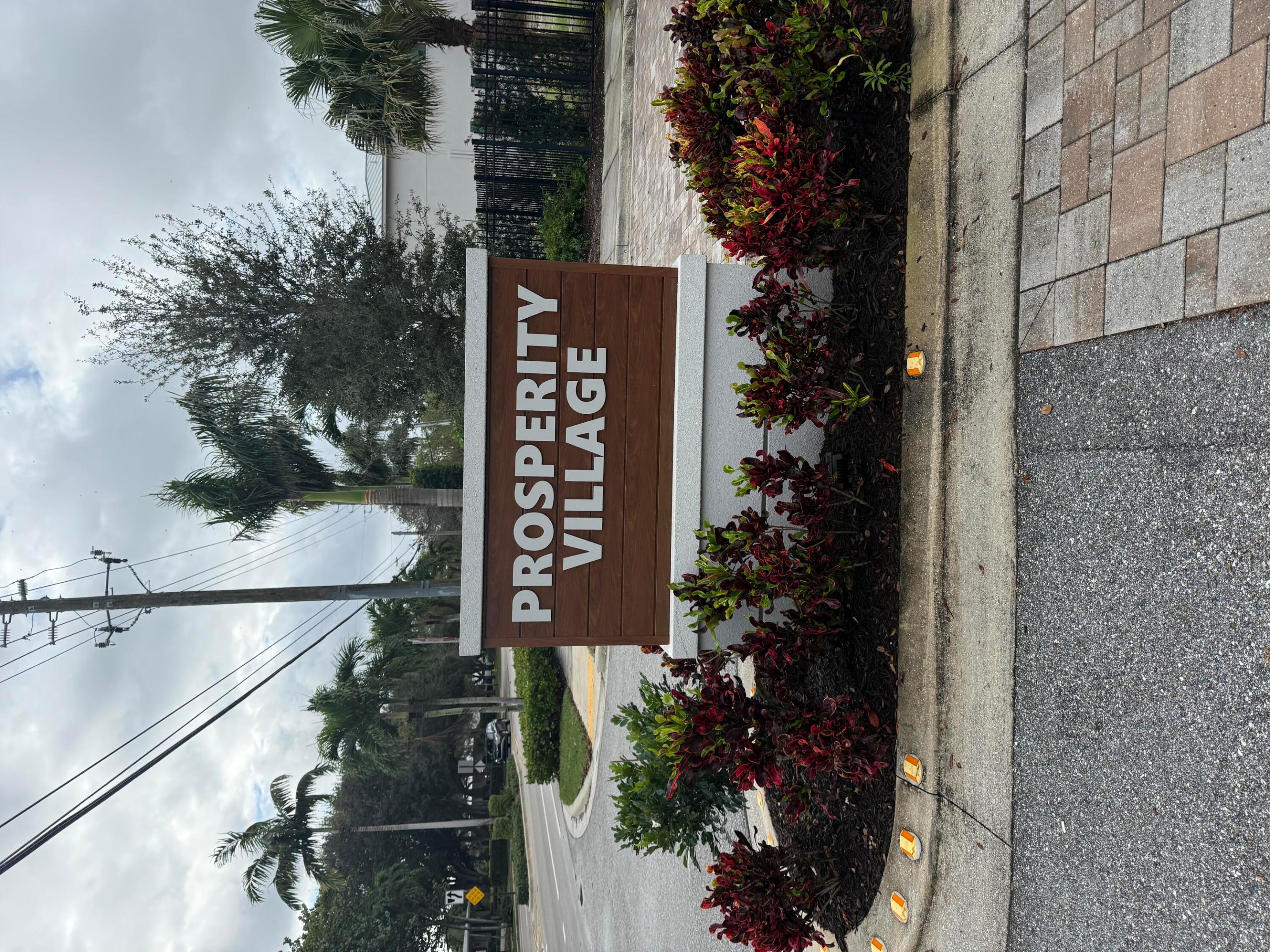 Prosperity Village entrance sign surrounded by shrubs beside a palm-lined street.