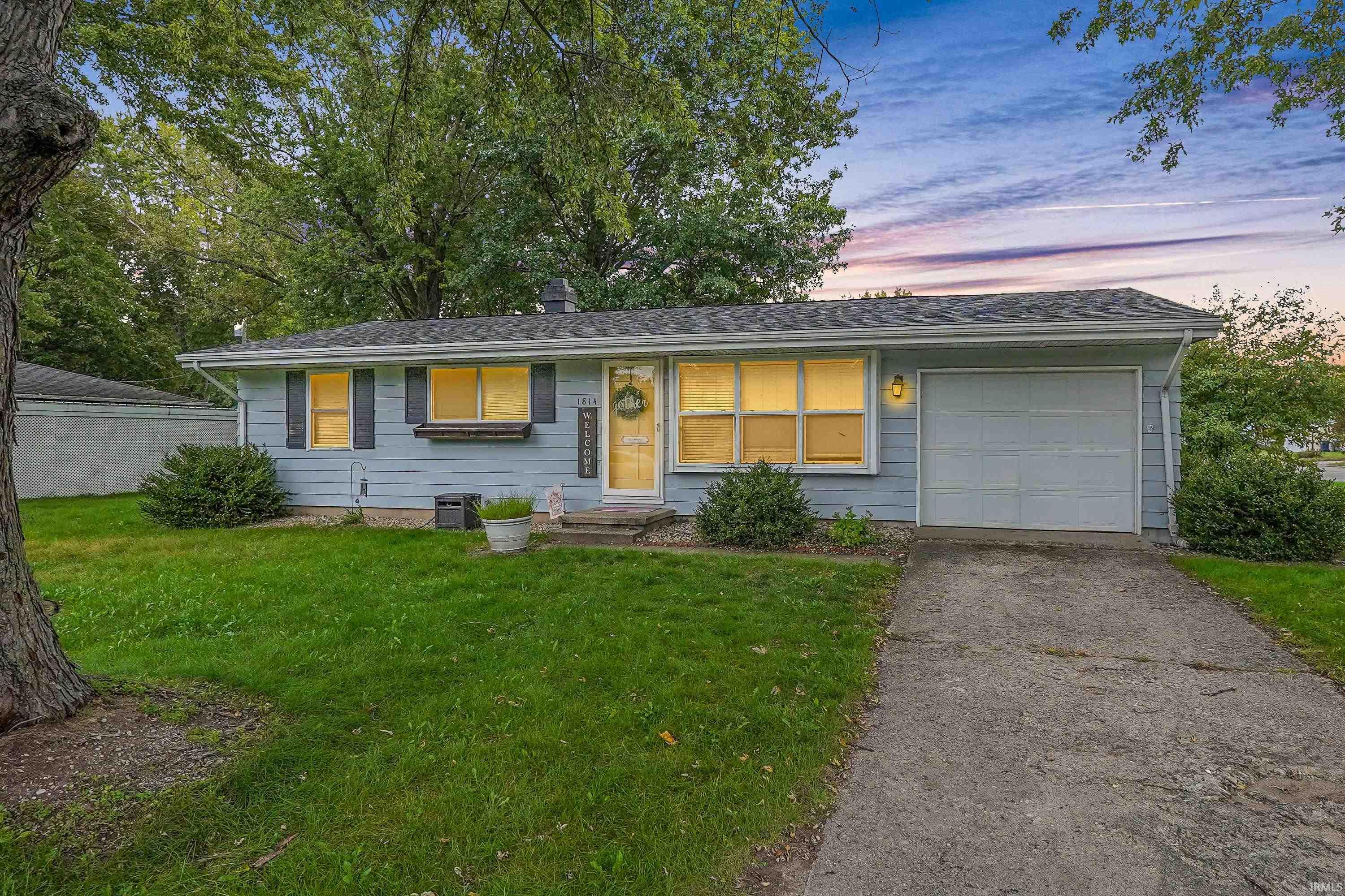 Light blue single-story home with a one-car garage, leafy yard, and warmly lit windows at dusk.