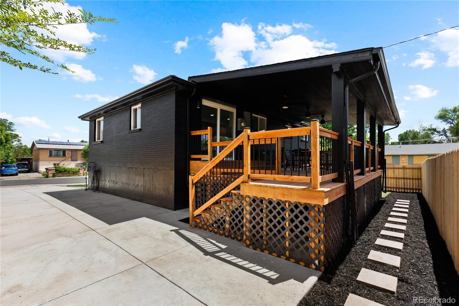 Dark brick house with a covered raised deck and wooden stairs beside a driveway and fenced path.