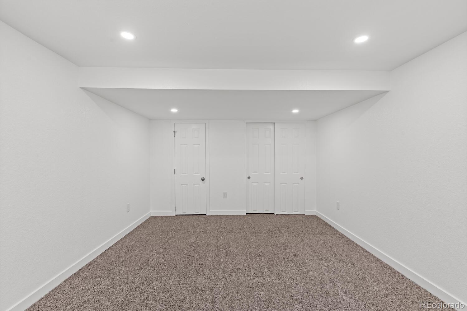 Empty carpeted room with white walls, recessed lights, and three white doors.