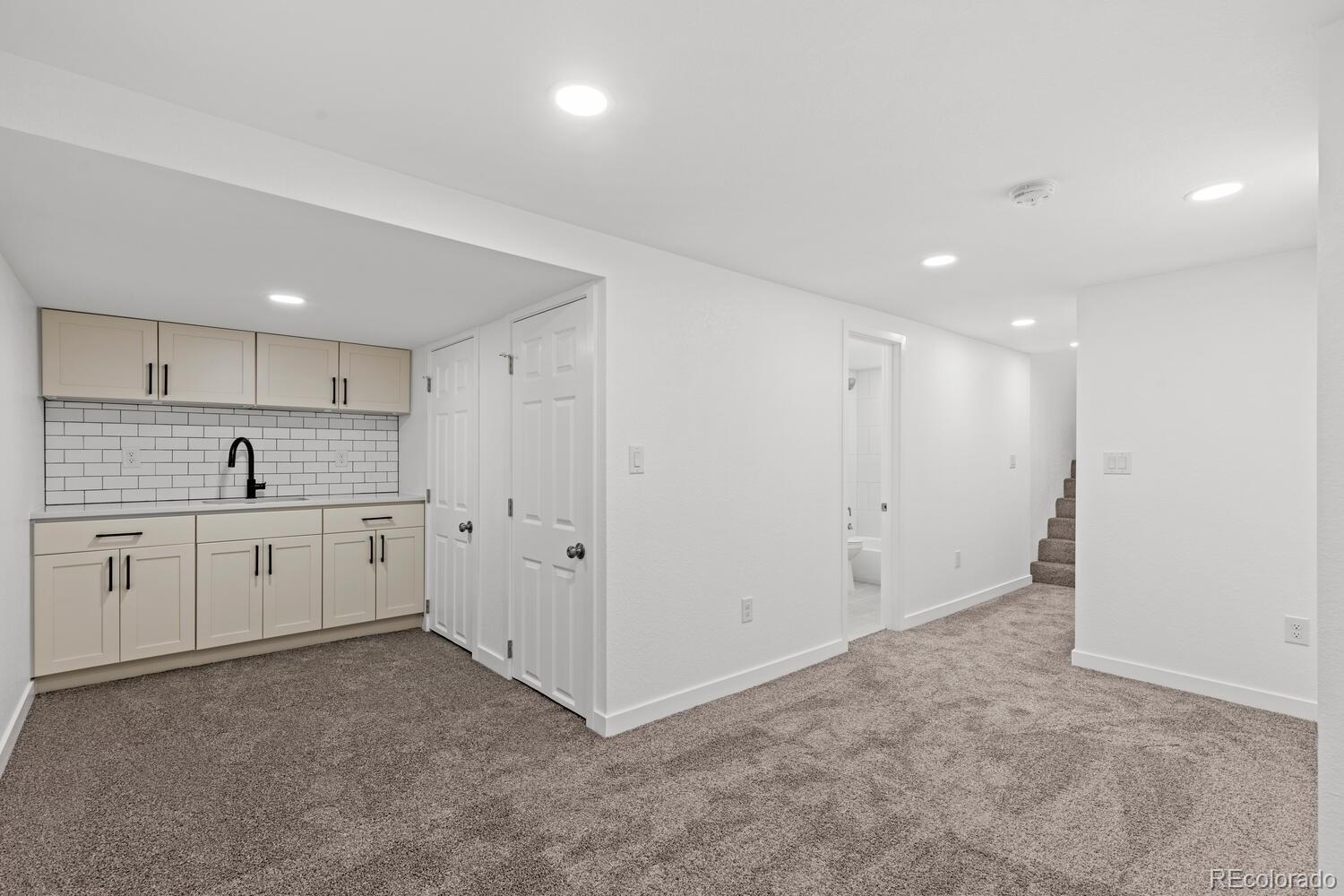 Finished basement with a compact kitchenette, carpeted open area, and stairs.