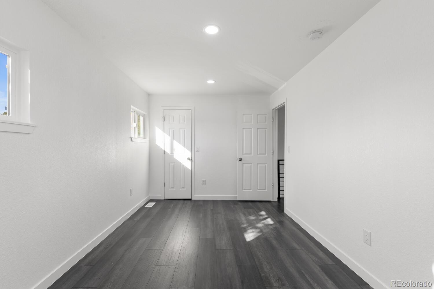 Empty room with dark plank flooring, two doors, and sunlight through small windows.