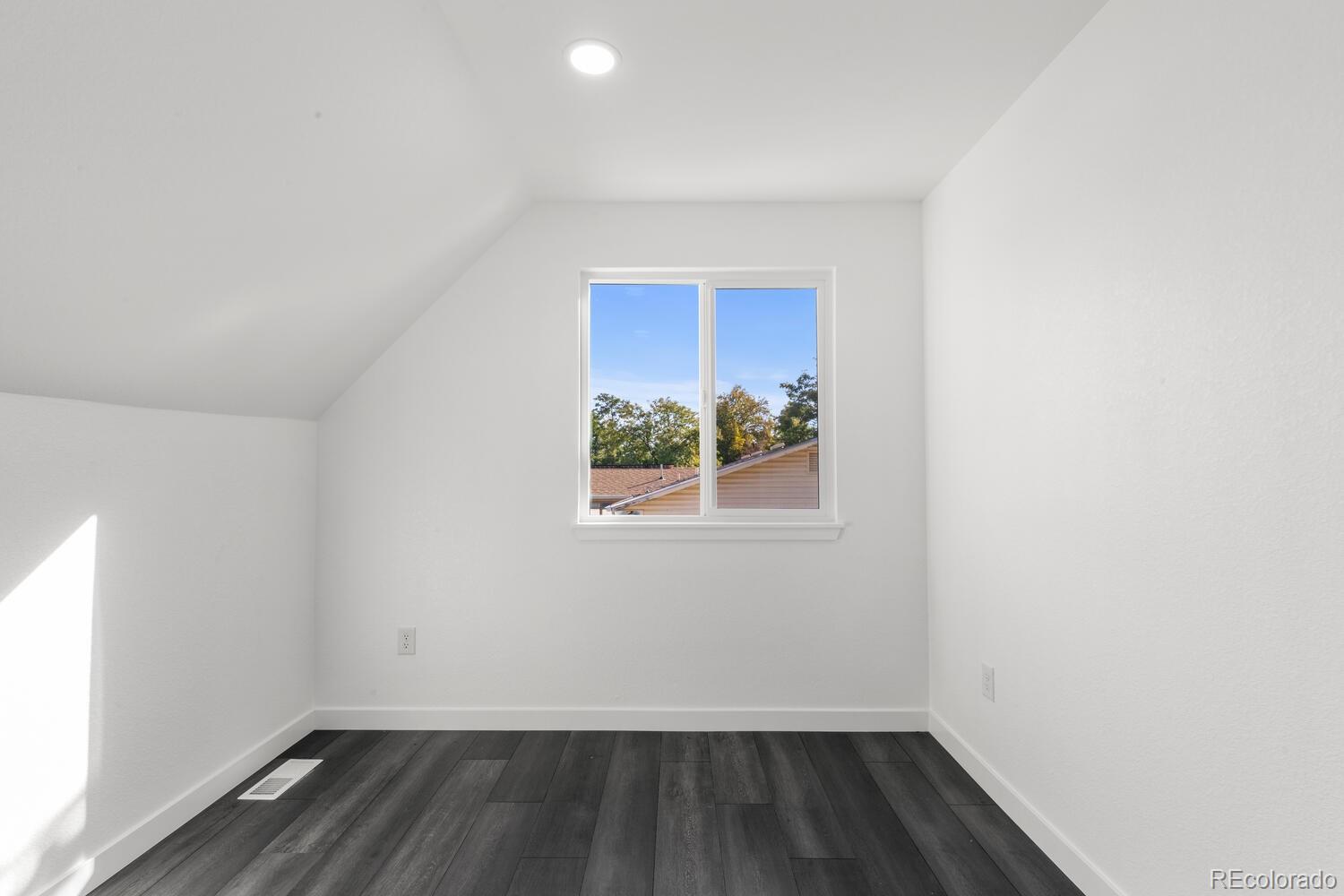 Empty bedroom with a sloped ceiling, dark plank flooring, and a window overlooking rooftops and trees.