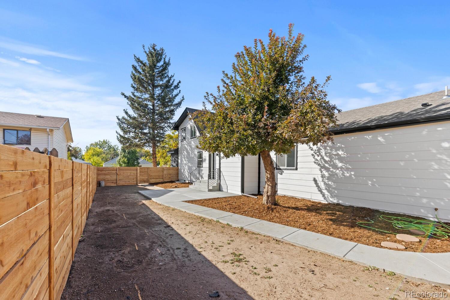Fenced backyard with a concrete walkway, mature trees, and mulched beds beside the house.