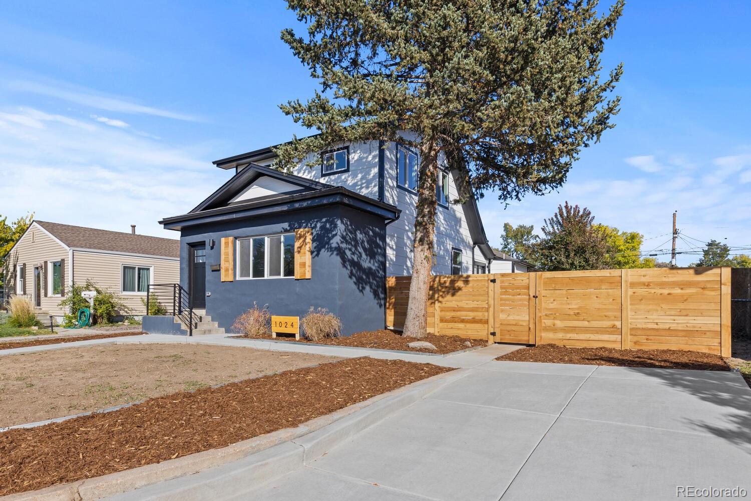 Two-story blue-gray home with wood shutters, a tall evergreen, and a wooden privacy fence.
