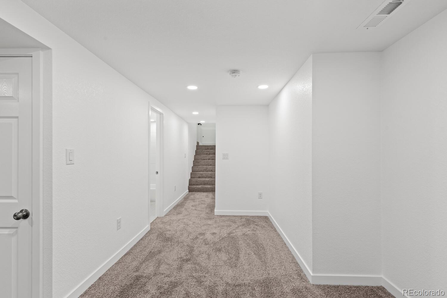 Empty carpeted hallway with white walls and stairs at the far end.