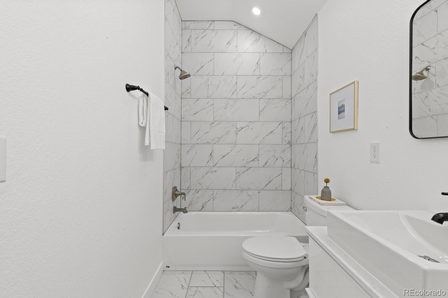 Bathroom with a marble-look tiled tub and shower, toilet, and white sink vanity.