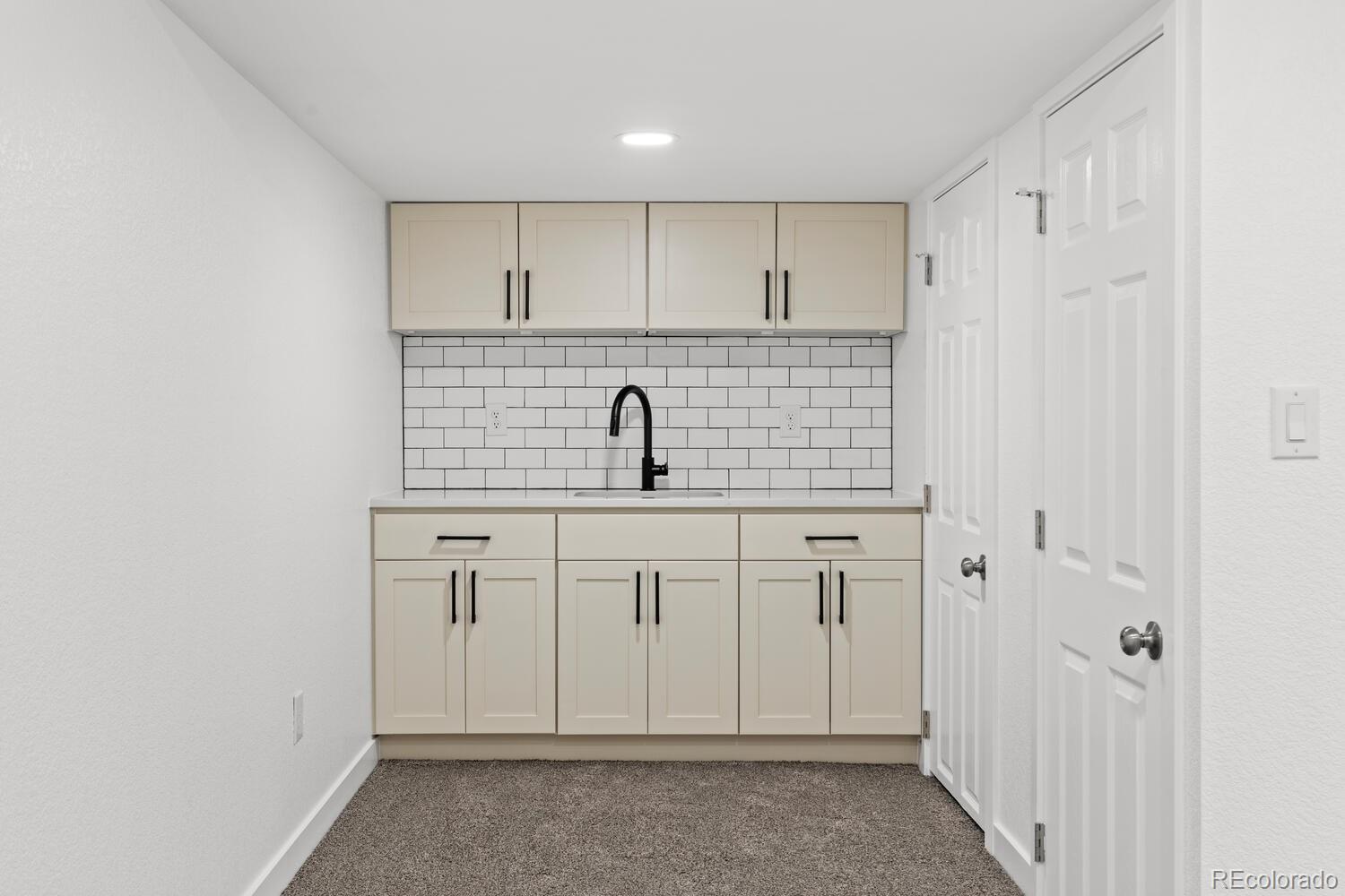 Compact kitchen with cream cabinets, subway-tile backsplash, black faucet, and gray carpet.