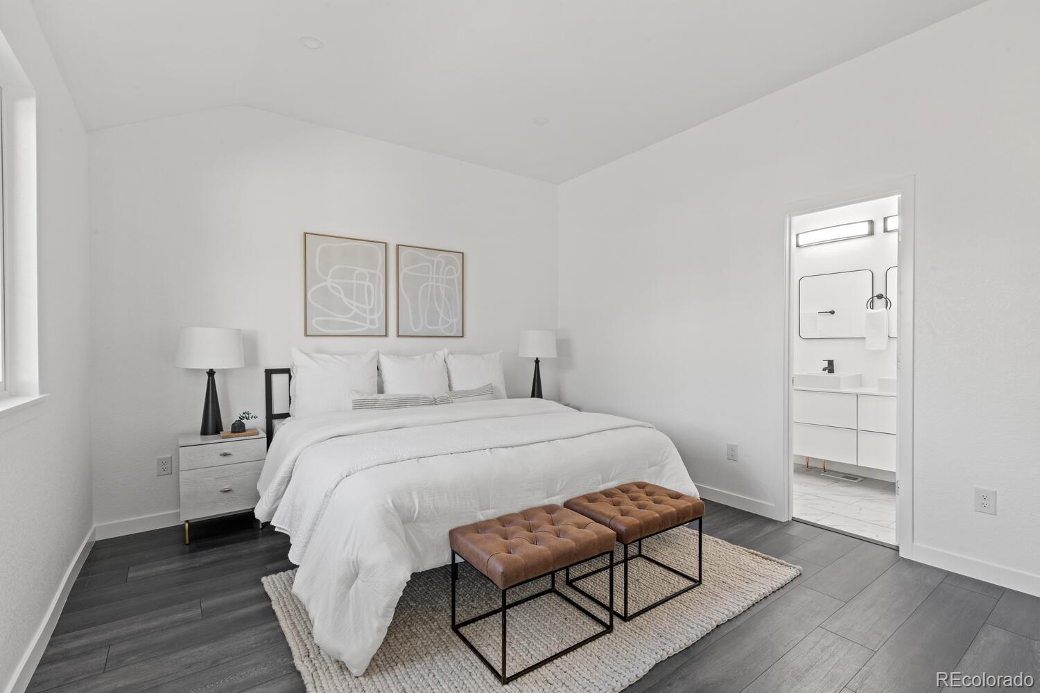 Staged bedroom with a white bed, brown tufted benches, and an ensuite bathroom visible through the doorway.