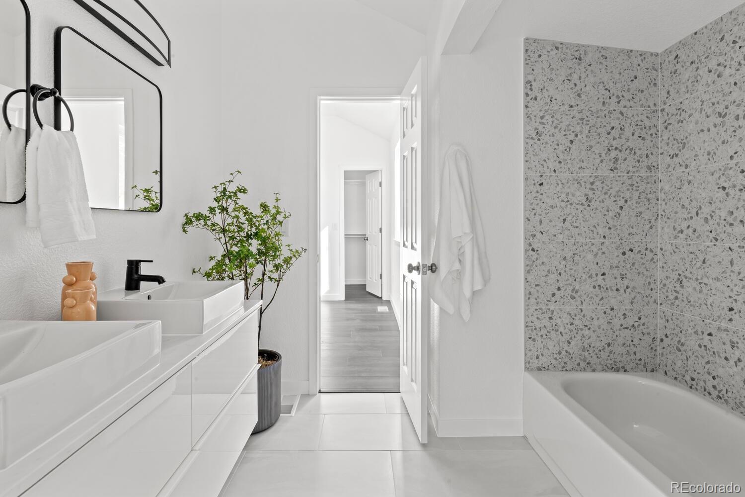 Bright bathroom with double vessel sinks, terrazzo-tiled tub surround, and black-framed mirror.