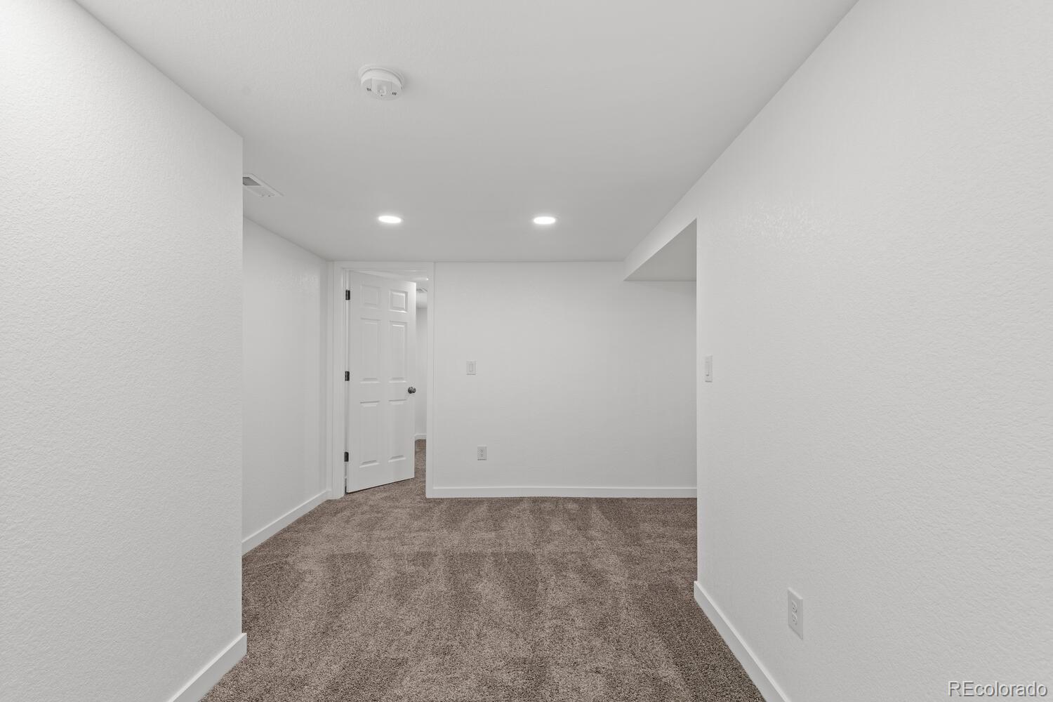Empty carpeted room with white walls, recessed lights, and an open door.