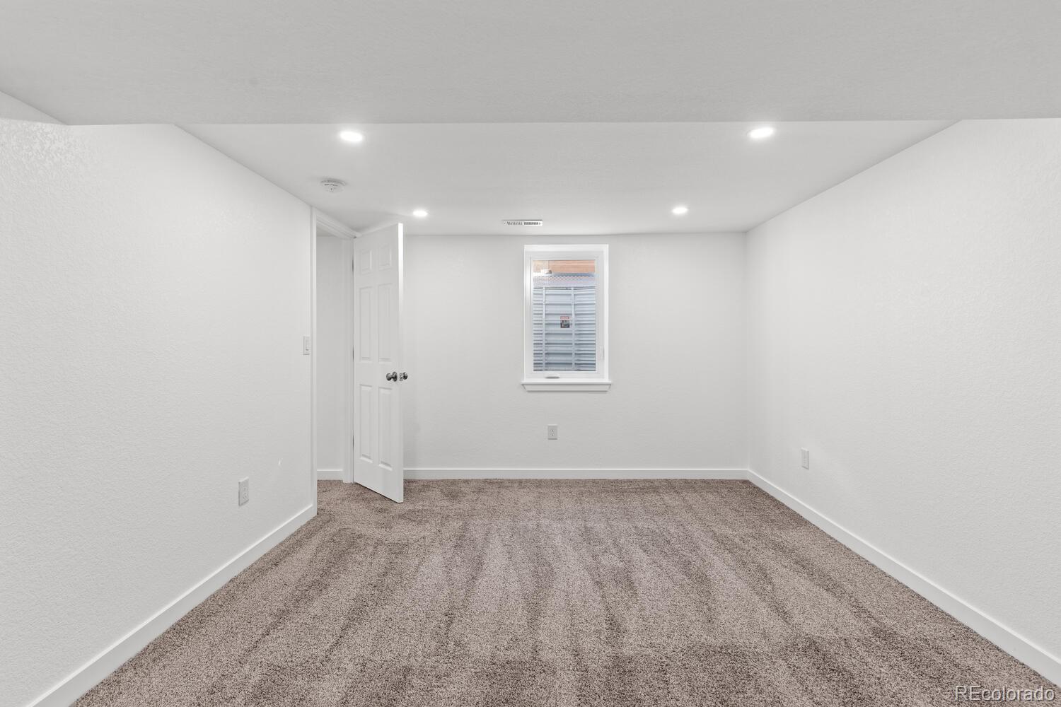 Empty carpeted room with white walls, recessed lights, a small window, and an open door.