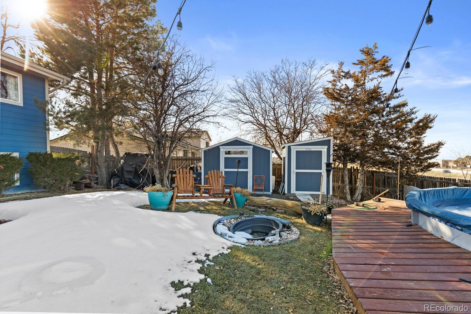 Snow-dusted backyard with two blue sheds, a wooden deck, fire pit, and covered pool.