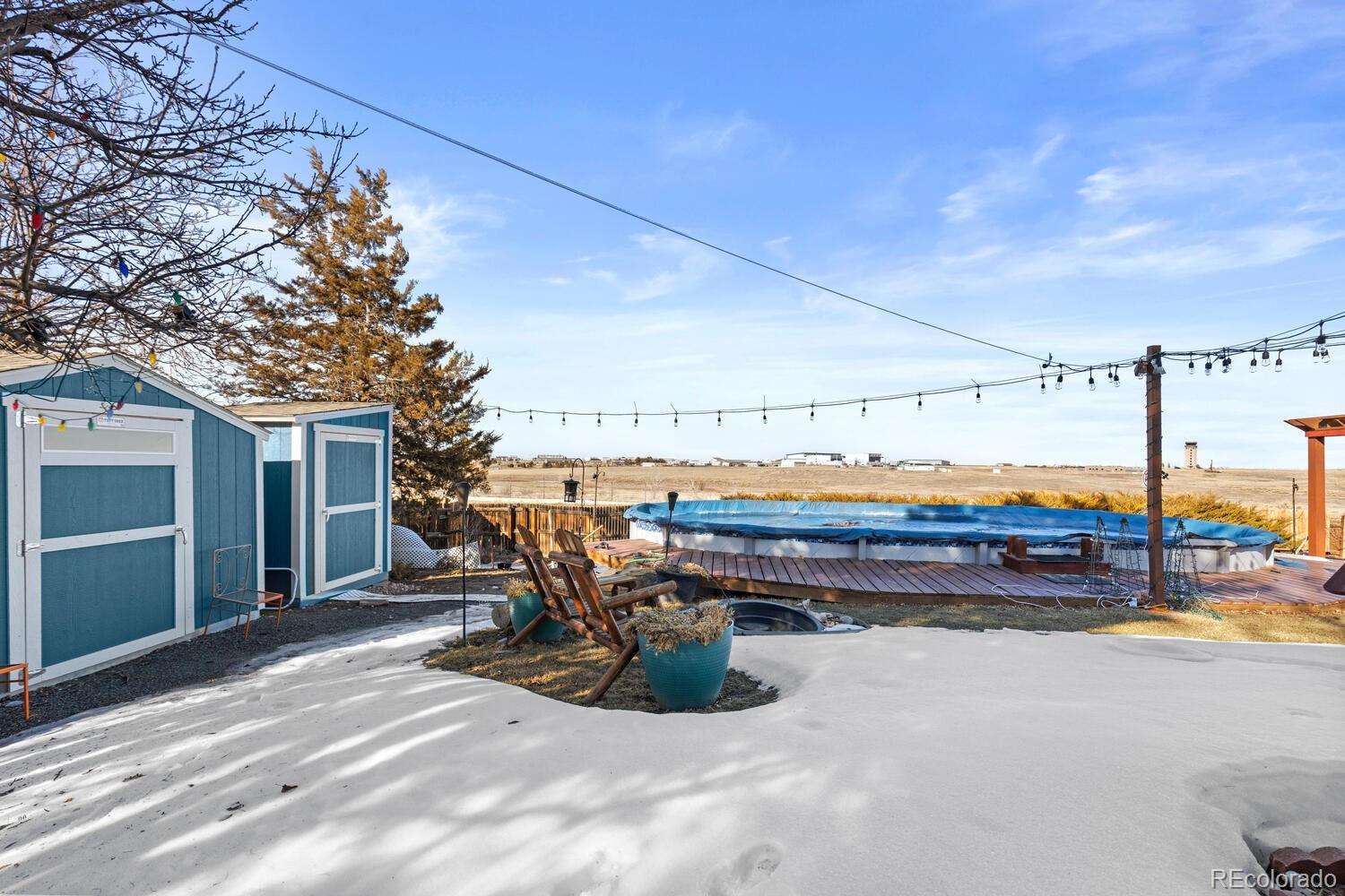 Backyard pool and deck with blue sheds, patio chairs, string lights, and snow.