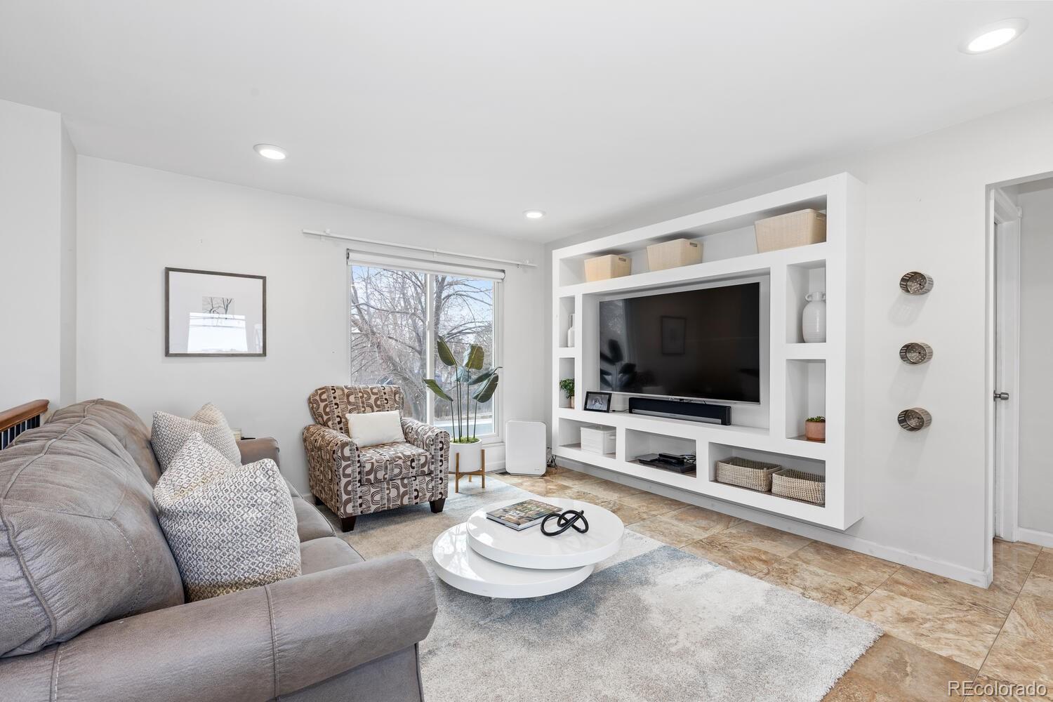Living room with built-in television shelving, gray sectional, patterned armchair, and a window overlooking bare trees.