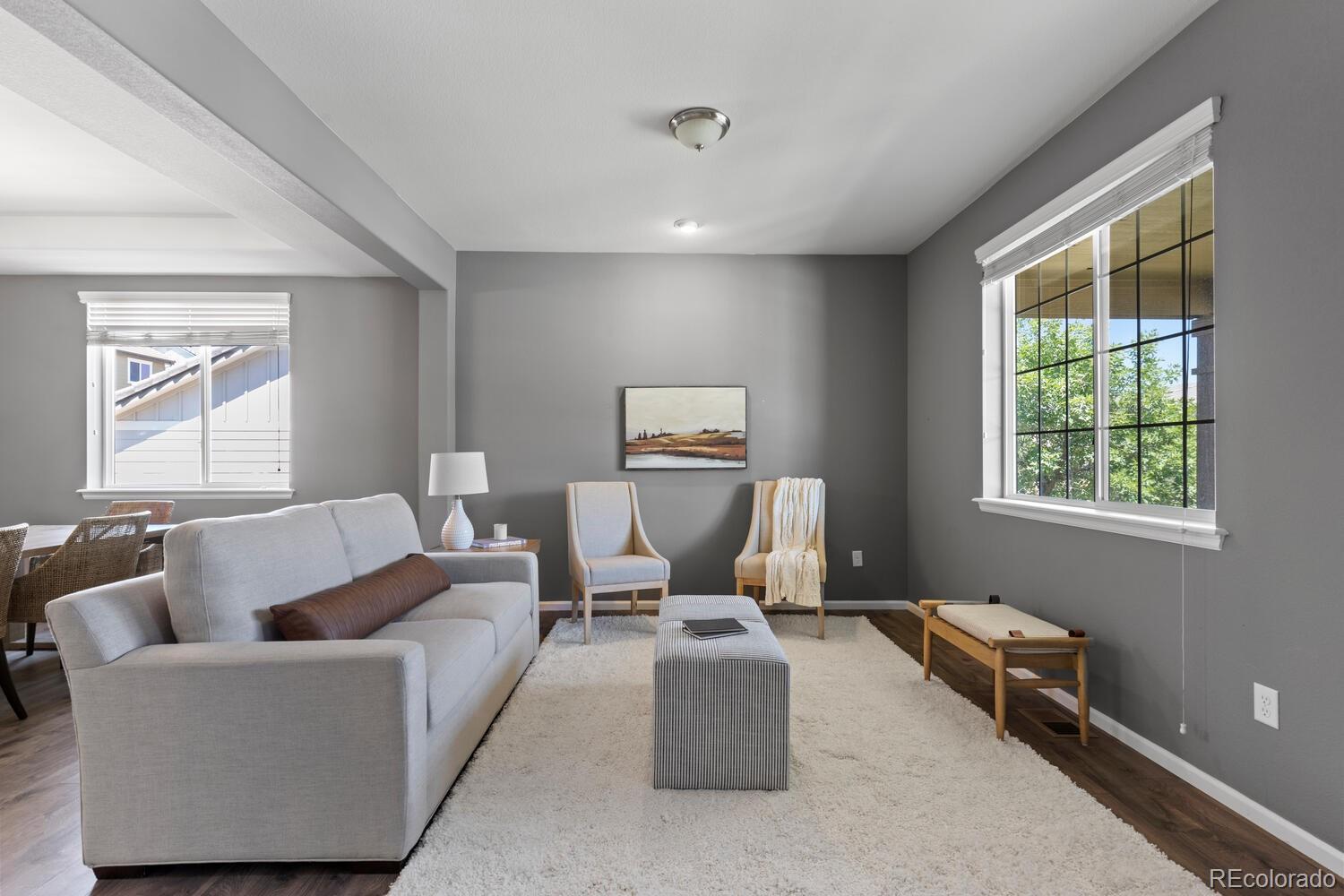 Staged living room with a light gray sofa, two accent chairs, and large sunlit windows.