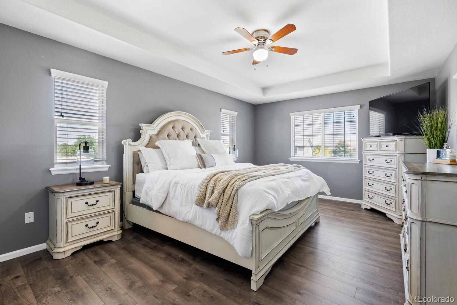 Furnished bedroom with a cream bed, gray walls, dark wood floors, and a wall-mounted television.