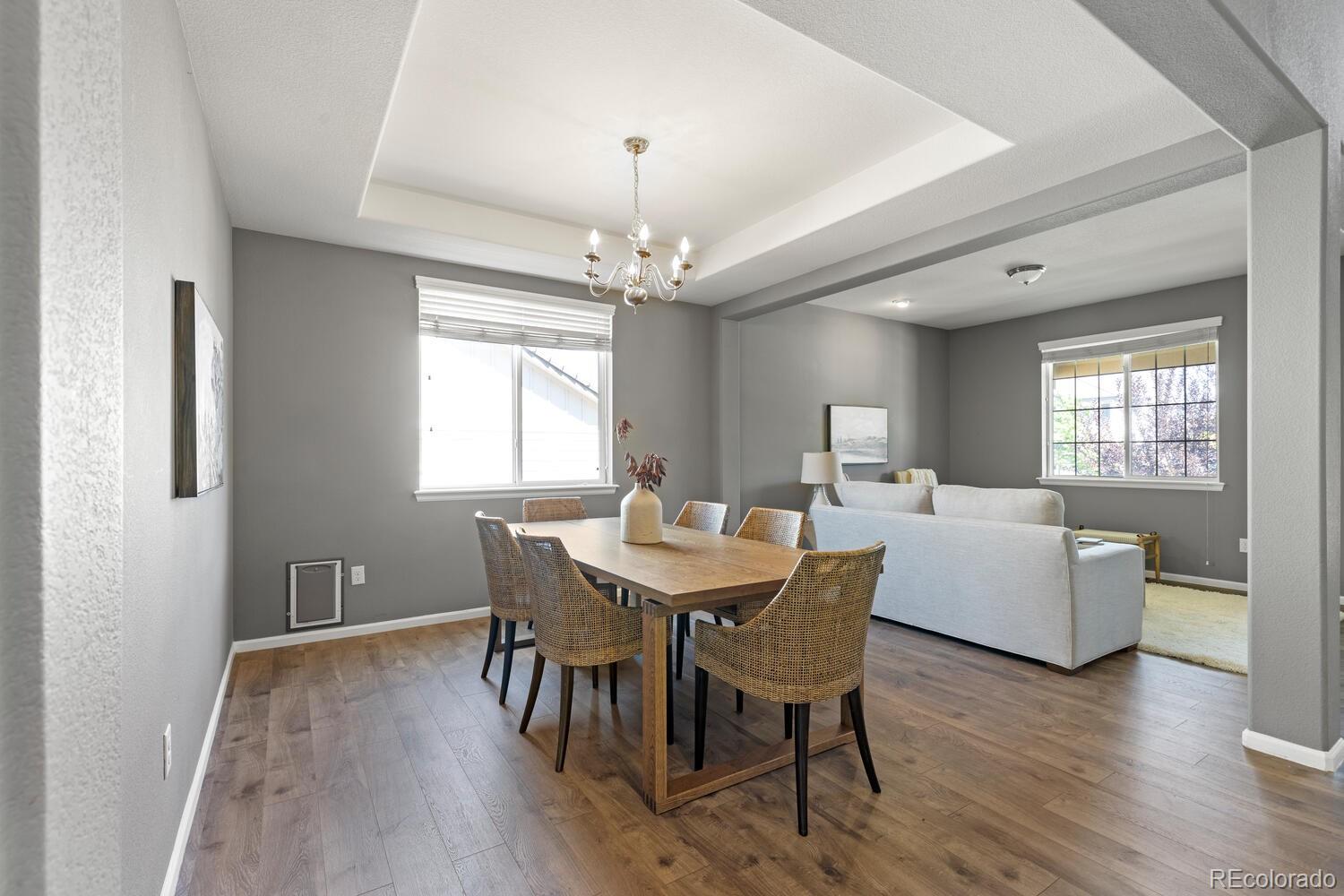 Dining and living area with a tray ceiling, chandelier, wooden table, woven chairs, and light sofa.