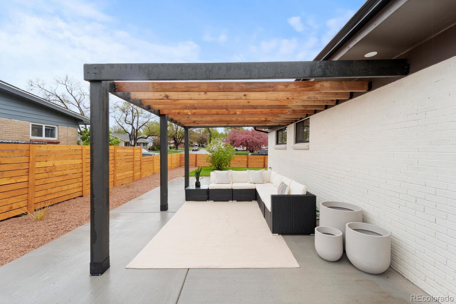 Covered patio with timber pergola, white-cushioned sectional, cream rug, and wooden privacy fence.