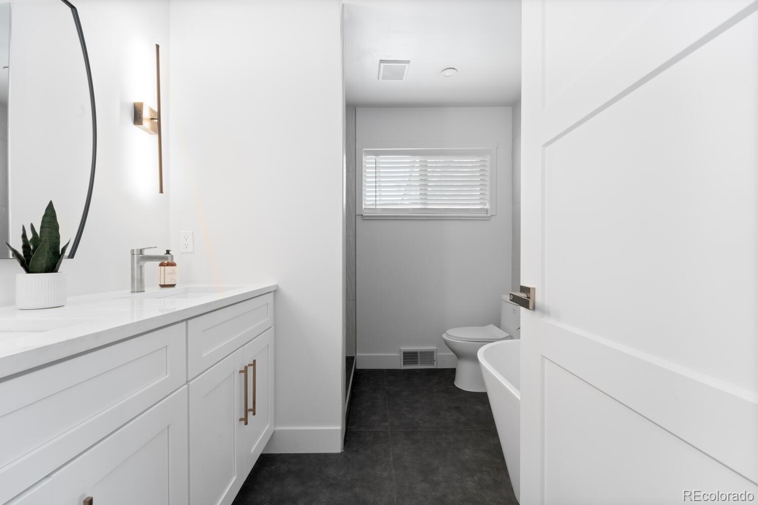 Bathroom with a white vanity, round mirror, dark tile floor, and window with blinds.