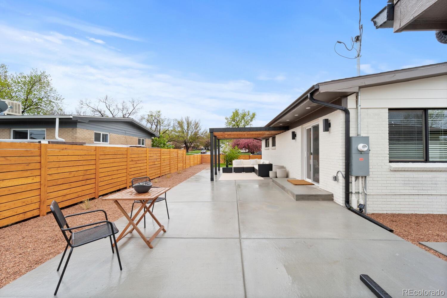 Concrete patio beside a white brick home, with a wood privacy fence, pergola, and outdoor seating.