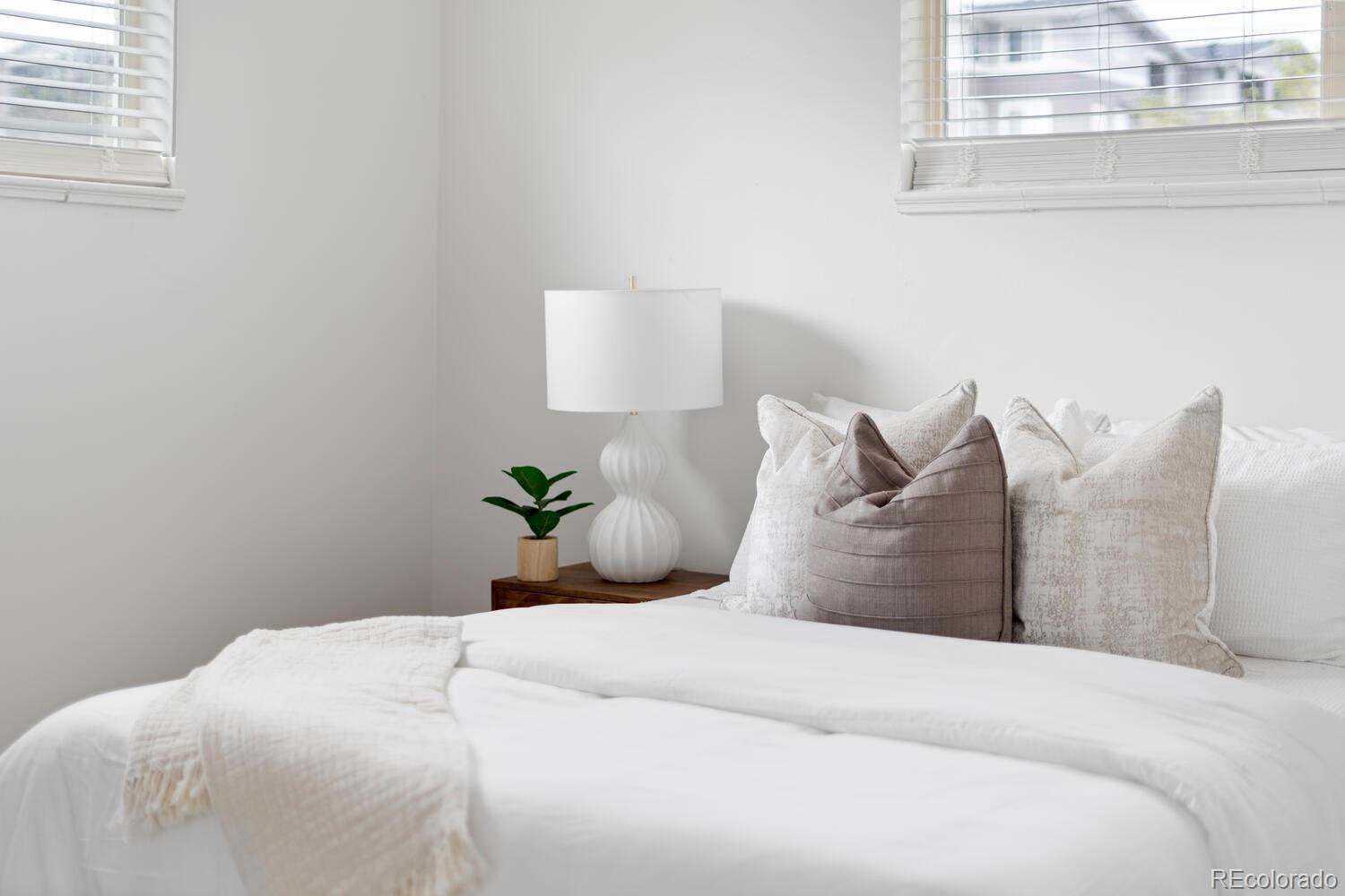 Staged bedroom with a white bed, taupe accent pillows, and a bedside table with a lamp and plant.