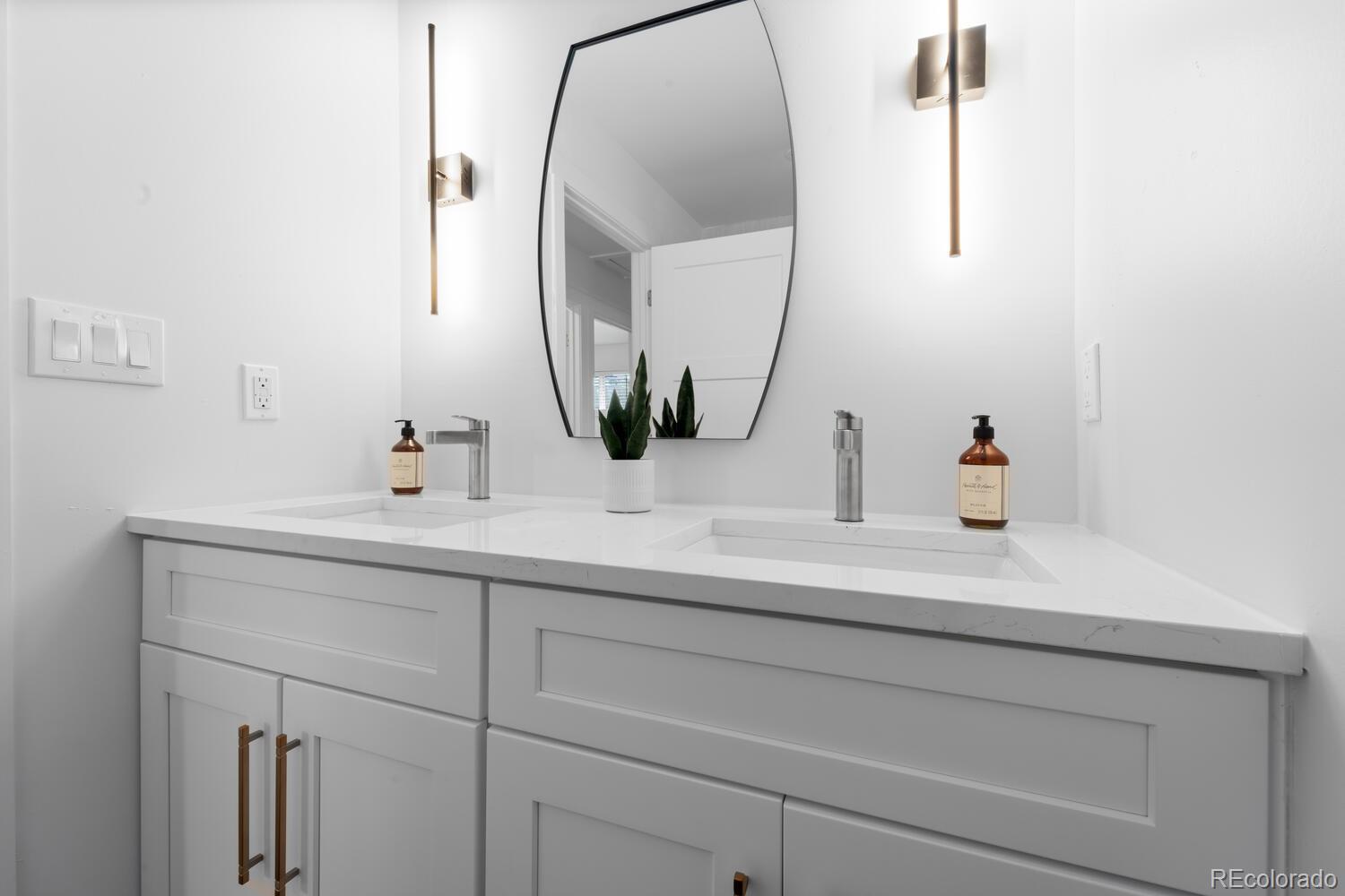 Bathroom double vanity with white cabinetry, an oval mirror, and brass wall lights.
