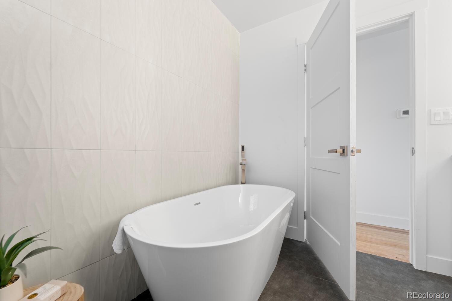 Modern bathroom with a freestanding white soaking tub and pale textured tile wall.