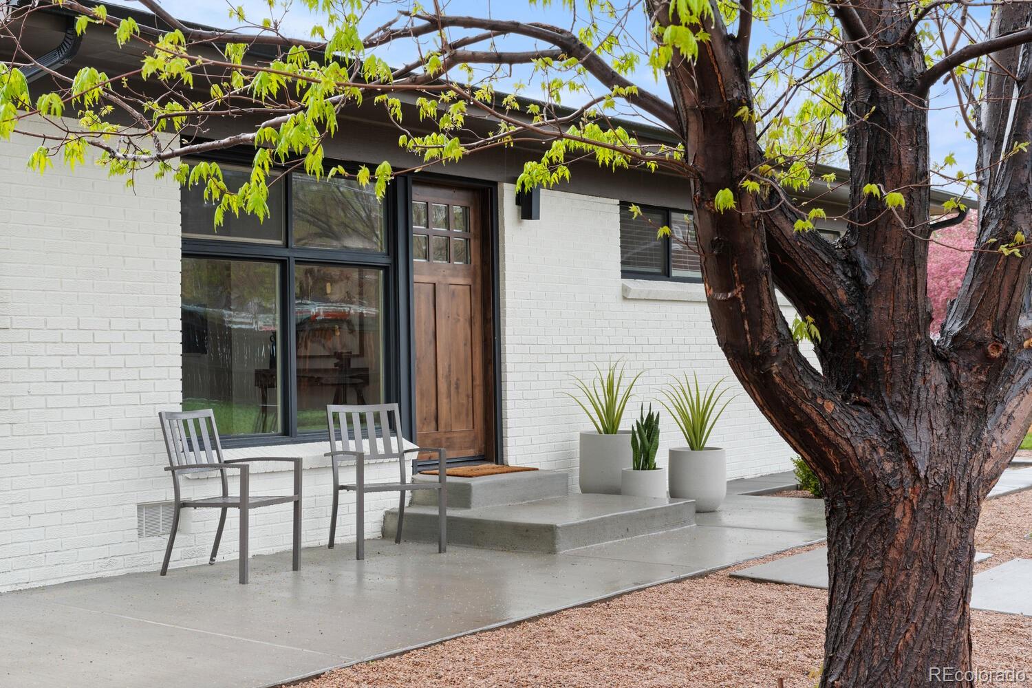 White-brick home entrance with a wooden door, patio chairs, and a large leafy tree.