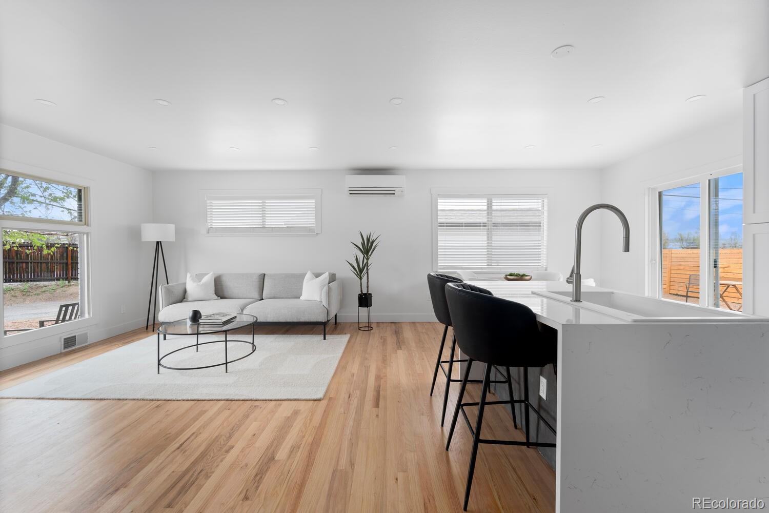Open-plan living room and kitchen with a gray sofa, hardwood floors, and a white island with black stools.