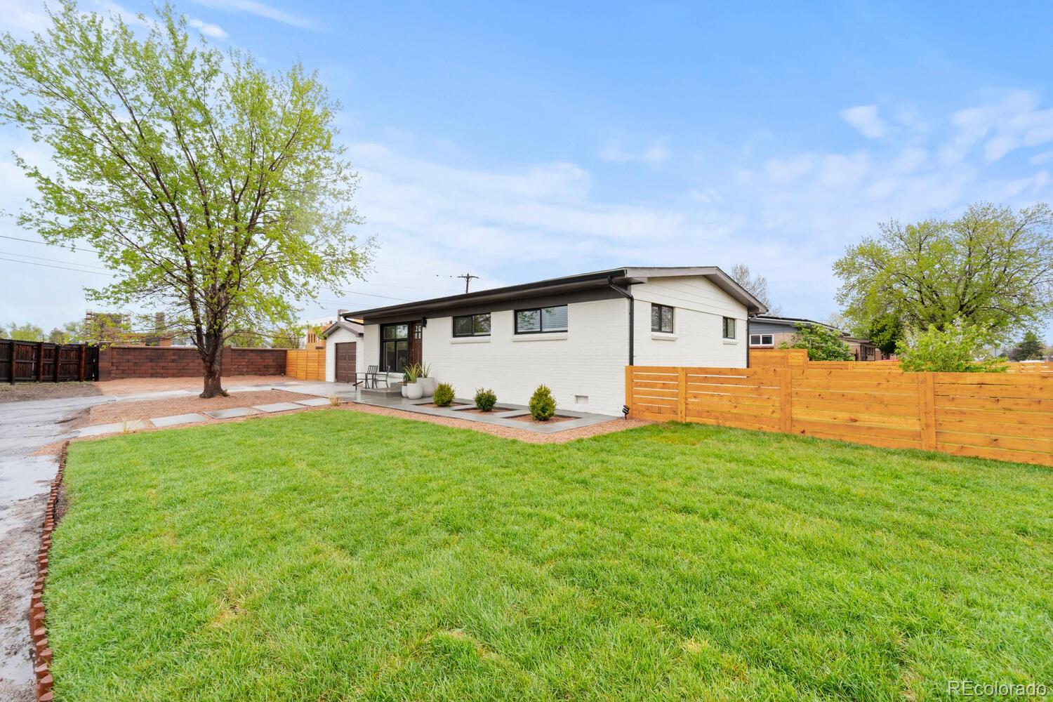Single-story white brick home behind a broad lawn, with wooden fencing and a small patio.