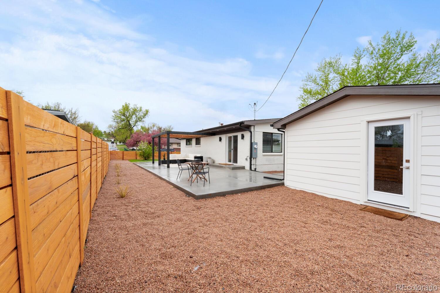 Backyard patio with pergola and seating beside a white home, enclosed by a tall wood fence.