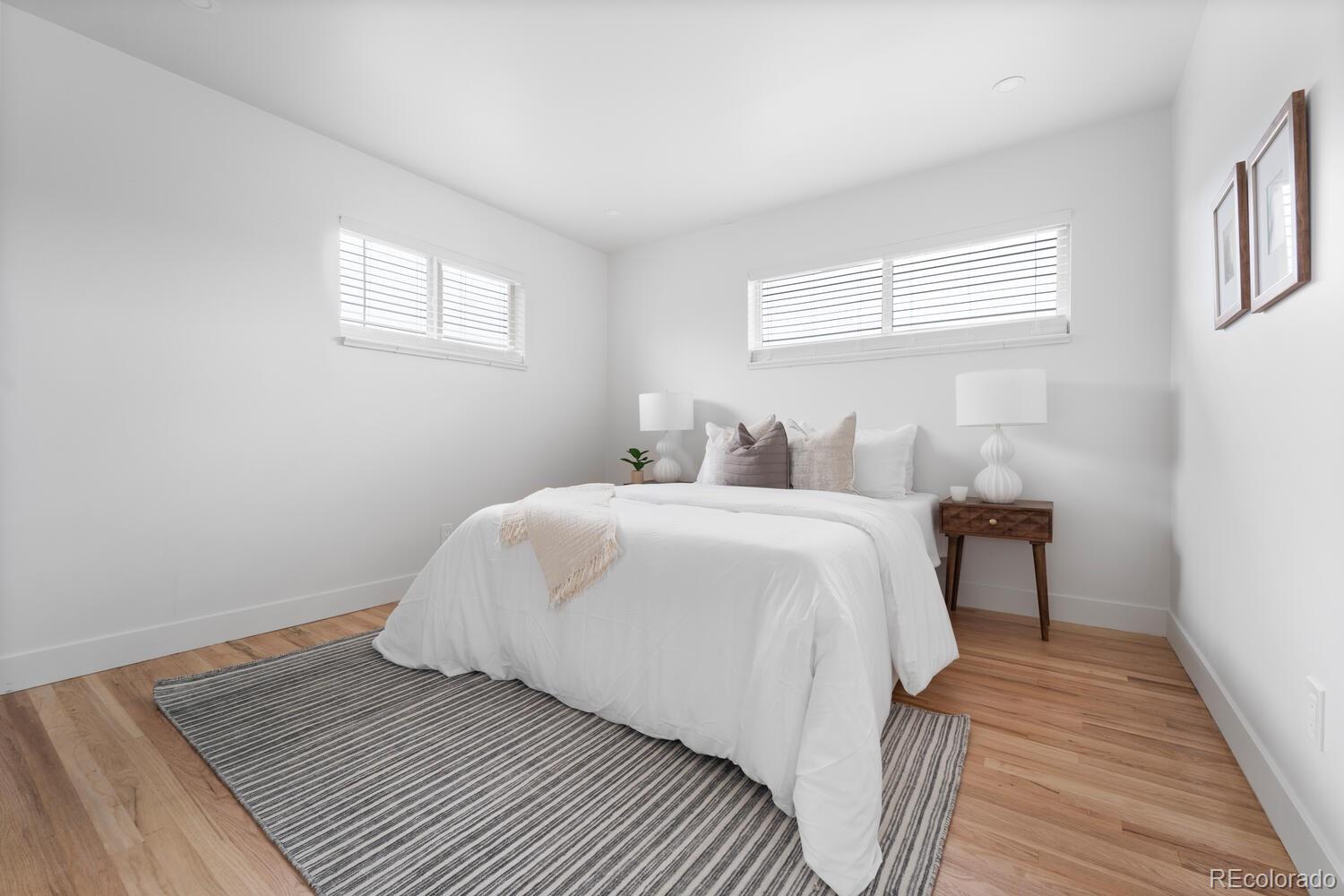 Staged bedroom with a white bed, striped rug, hardwood floors, and high windows with blinds.