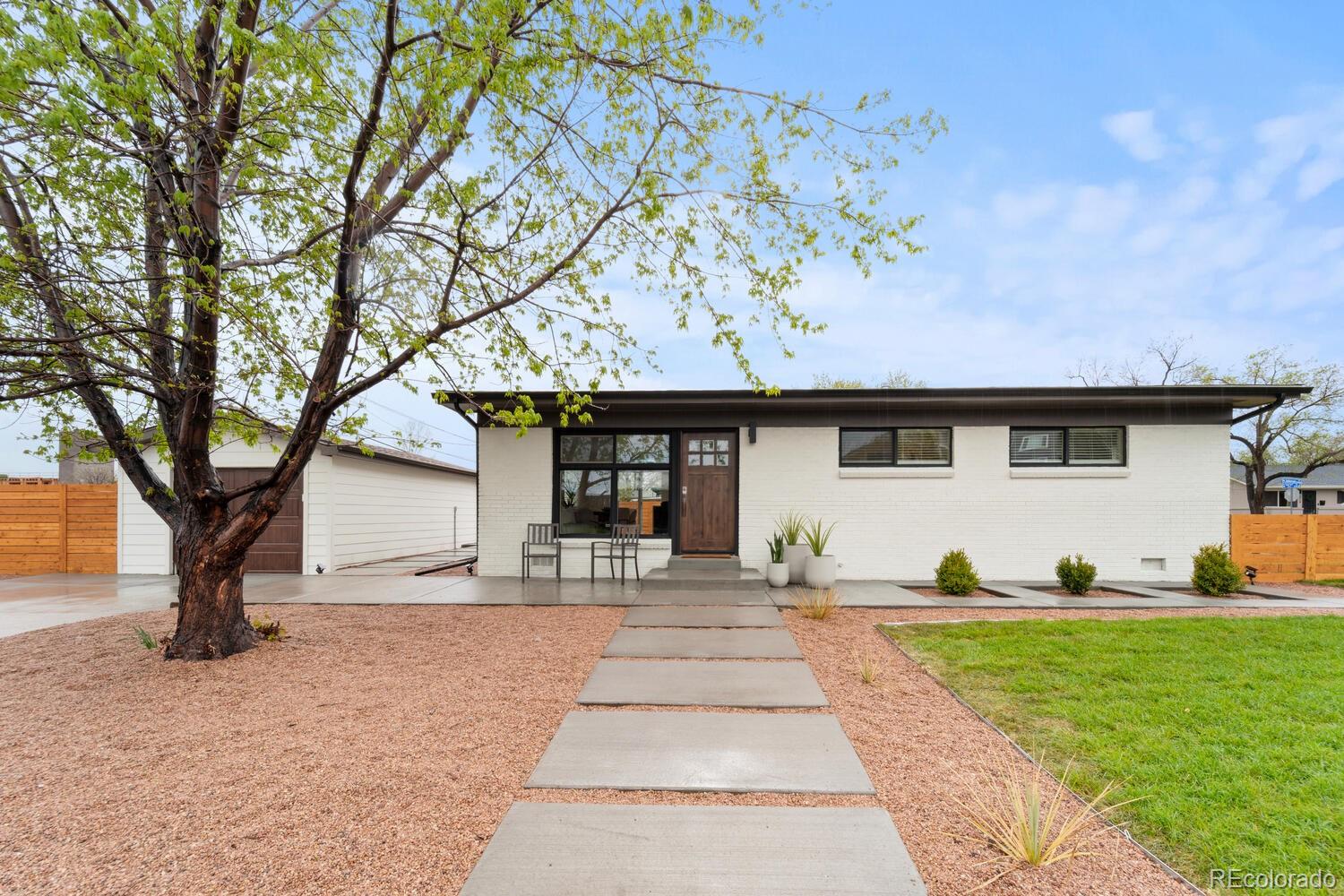 Single-story white brick home with a wood entry door, concrete walkway, and mature tree.